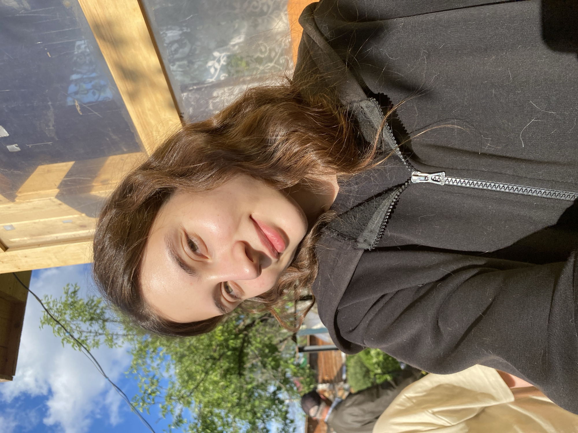 A young woman is posing for a selfie outdoors. She is wearing a black hooded sweatshirt, partially zipped. Her hair is brown and wavy. The background includes trees, sky with clouds, and a wooden structure. The location and city are not identifiable in the image. There are people behind her.
