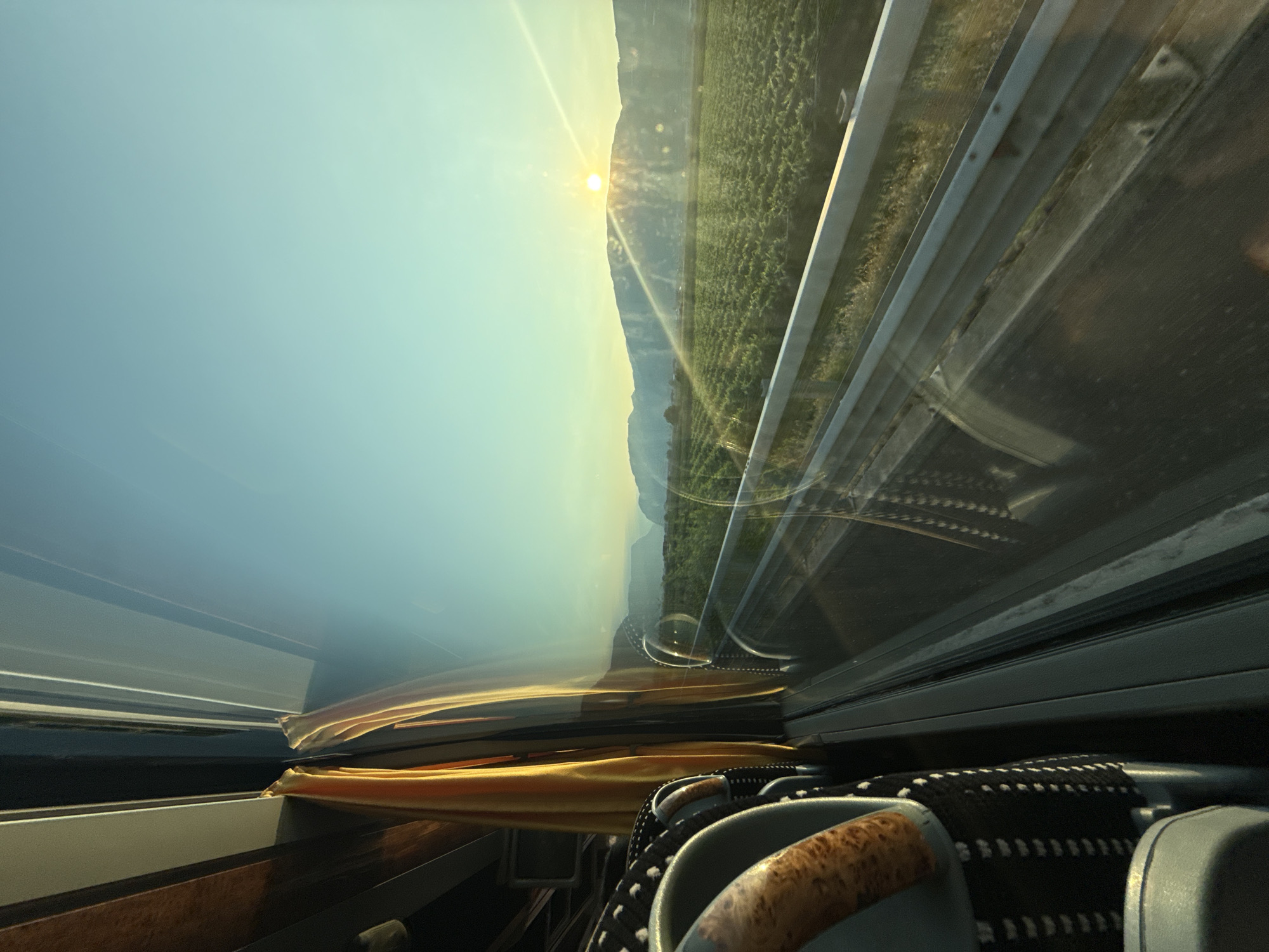 The image shows a view from the window of a vehicle, likely a bus, traveling along a highway. Inside the vehicle, a yellow curtain is drawn back, revealing a seat with a patterned upholstery and a wooden armrest. Outside the window, a vibrant green field stretches towards a line of mountains in the distance. The sun is low in the sky, casting a warm glow and a lens flare across the scene, suggesting either sunrise or sunset. A metal guardrail is visible along the side of the road. The sky is a clear, gradient blue, transitioning to a lighter hue near the horizon. The overall impression is one of serene travel through a rural, mountainous landscape. There is no visible indication of the city of Zambana, Italy, within the image itself.