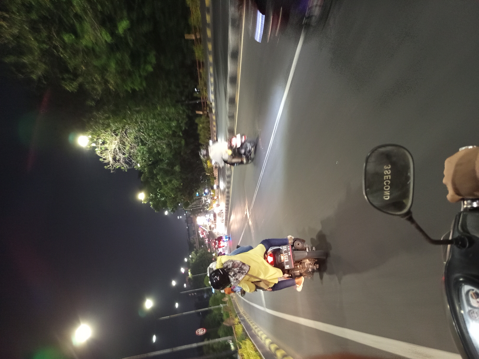The image depicts a night scene on a multi-lane road in Buduran, Indonesia. In the foreground, a motorcycle with two occupants is moving forward. The rider is wearing a helmet, and the passenger, positioned behind the rider, is wearing a yellow shirt and a headscarf. The motorcycle has a license plate visible that reads "N 5731 XB". The rider's left hand is on the handlebars, and a hand with a ring is also visible on the right side, indicating this is likely a first-person perspective from a motorcycle.
The road surface is asphalt, with clear white lane markings. Streetlights are illuminating the scene from overhead poles, casting a bright glow and creating reflections on the wet road. Trees line the side of the road, with some of their foliage illuminated by the streetlights. In the distance, other vehicles and traffic lights are visible, suggesting ongoing traffic flow. A traffic sign with a red circle and diagonal line is present on the left side of the road, near a yellow barrier. The overall atmosphere is that of a busy urban road at night.