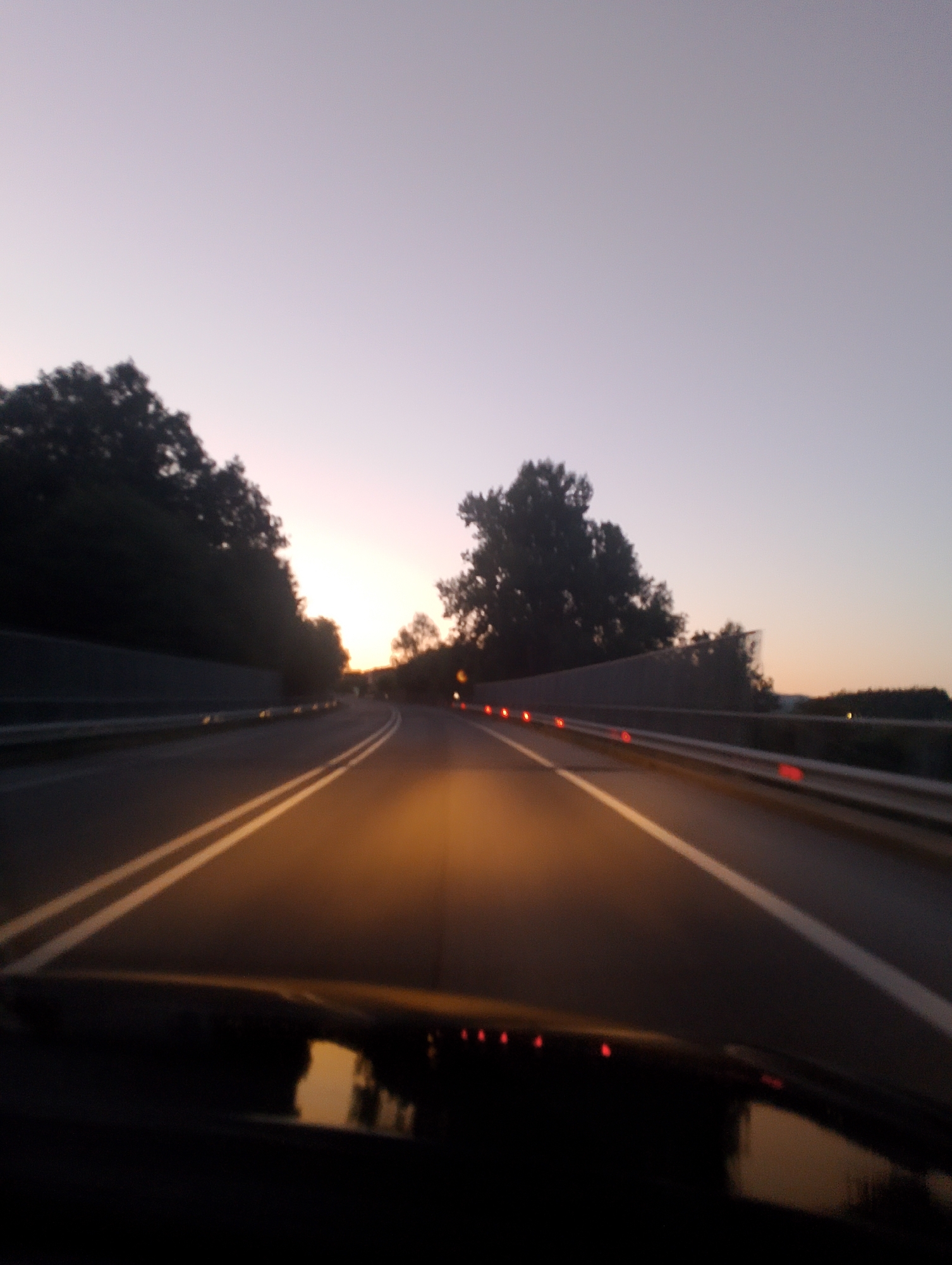 The image shows a perspective from inside a vehicle driving on a highway at dusk. The road has two white lines marking the lanes, and the sky is a pale gradient of purple and pink, indicating either sunrise or sunset. Trees line the sides of the road, casting dark shadows. Reflective markers are visible along the guardrail. The scene appears to be a rural or semi-rural highway, not a city location. It is impossible to determine the exact location within Borgo San Pietro, Italy, from this image. There are no people or identifiable landmarks present.
