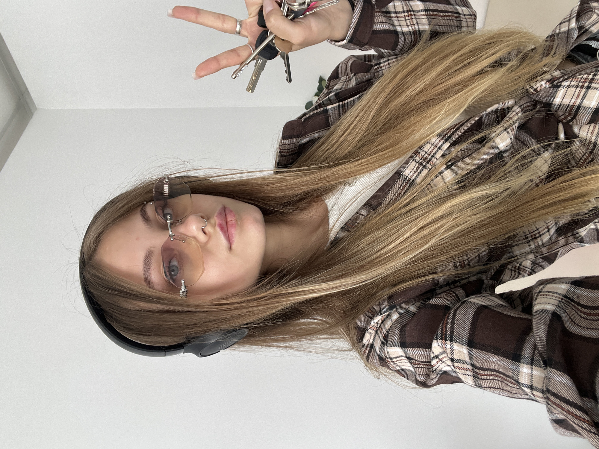 A fair-skinned person with long, light-brown hair, wearing rimless glasses, a nose ring, and black headphones, is holding keys in their right hand and making a peace sign. The person is also wearing a brown and beige plaid shirt. The background is a plain white wall. The location is not discernible from the provided image.