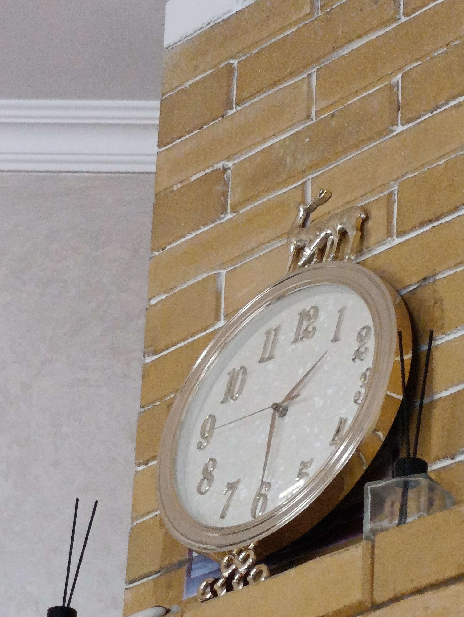 A close-up, eye-level shot shows a wall clock on a textured, light brown brick wall. The clock face is white with silver numerals and hands. A small, silver deer figurine is perched on top of the clock. To the right of the clock, a small, clear glass bottle with a black stopper and dark reeds is visible, suggesting it is an air freshener or diffuser. The background to the left of the brick wall is a textured, light grey surface with white decorative trim. The image is focused on the clock and its immediate surroundings. The scene is indoors and appears to be a domestic or decorative setting. The location of Qiblay, Tajikistan, is not determinable from the visual information presented in the image.