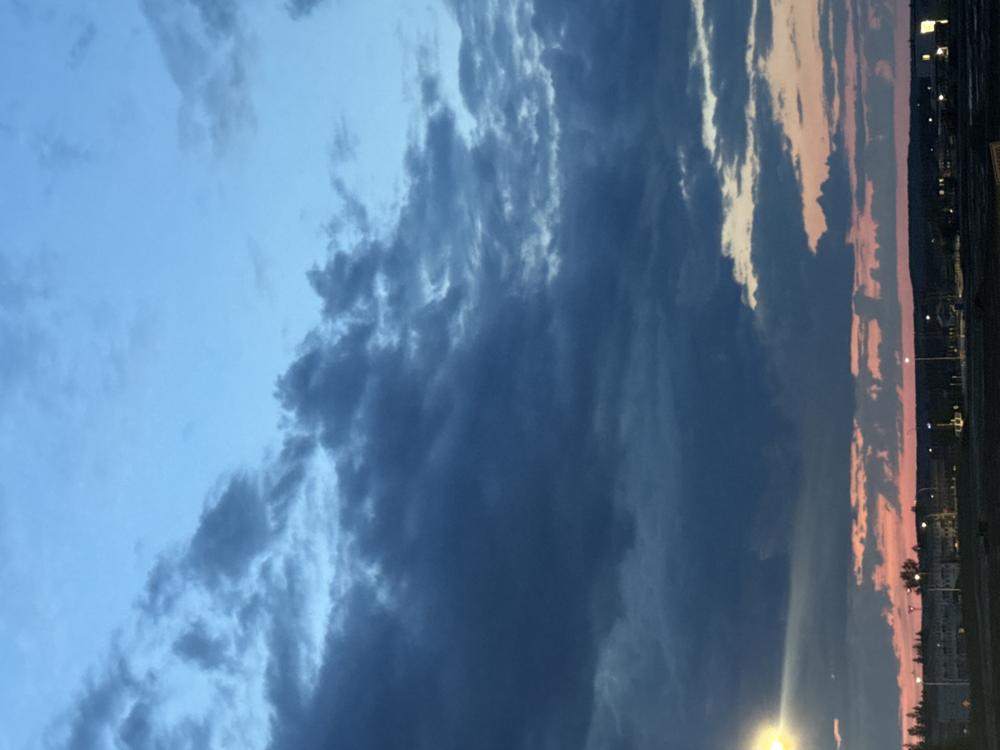 A low-angle, vertical, outdoor, eye-level photograph captures a wide expanse of the sky over a residential area at dusk. The sky transitions from a deep blue at the top to a hazy pink and orange near the horizon. Dark, heavy clouds dominate the upper and mid-sections of the frame, obscuring much of the lighter sky. Below the clouds, a faint, glowing light emanates from the horizon. In the lower portion of the image, a silhouetted landscape features the outlines of buildings, trees, and streetlights. A bright yellow light source, possibly a streetlight, is visible on the left side of the frame, casting a beam across the dark clouds. The scene appears to be located in Stony Plain, Canada, given the presence of typical Canadian residential architecture and vegetation under a vast sky. There are no people or animals visible in the frame.