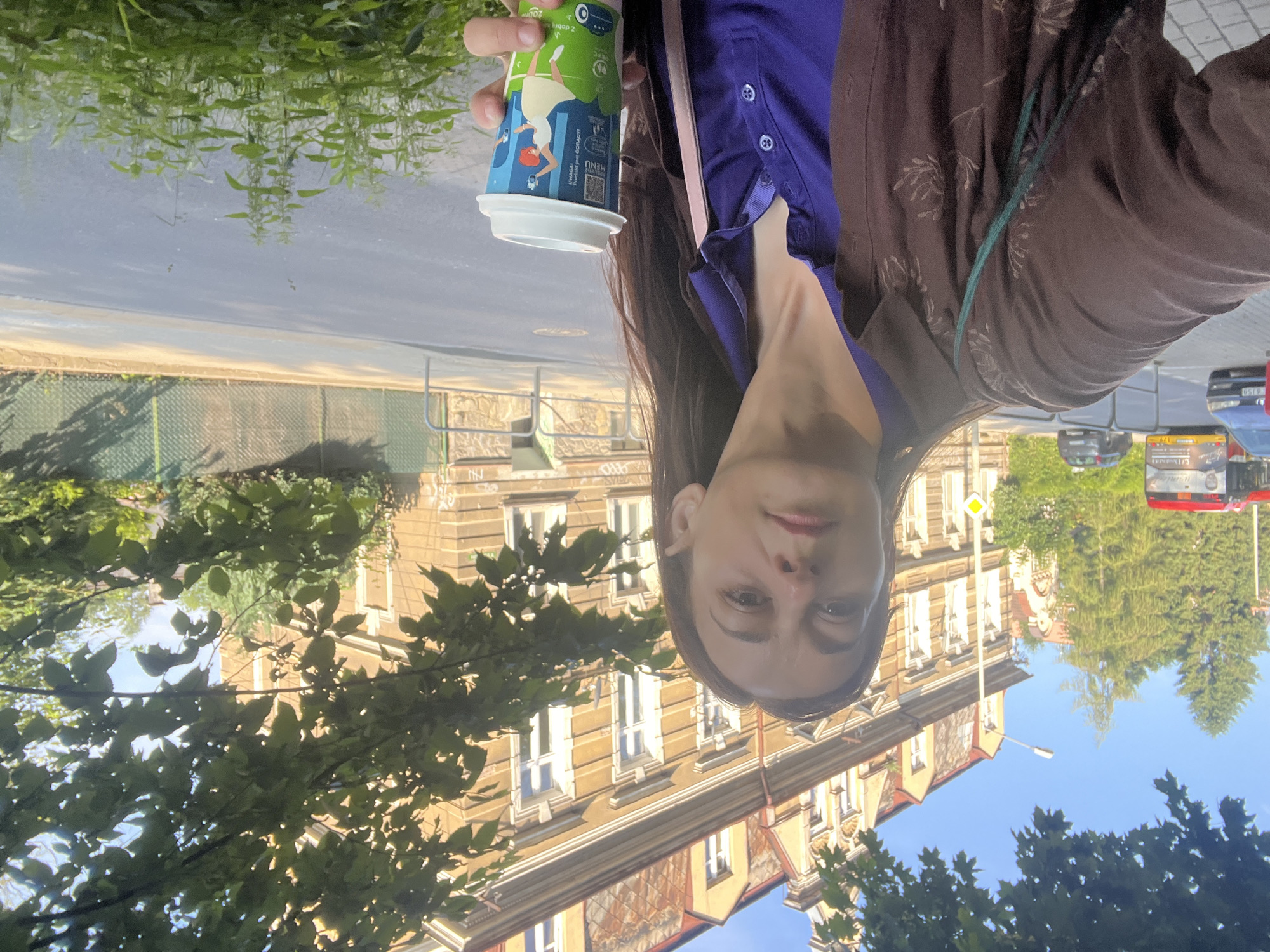 A woman stands on a sidewalk in front of an old, tan building, holding a coffee cup. She is wearing a brown patterned shirt over a purple collared shirt. The cup is decorated with a cartoon of a woman in a yellow dress and red hair. Behind her, trees and a street with a bus and cars are visible. The sky is blue.