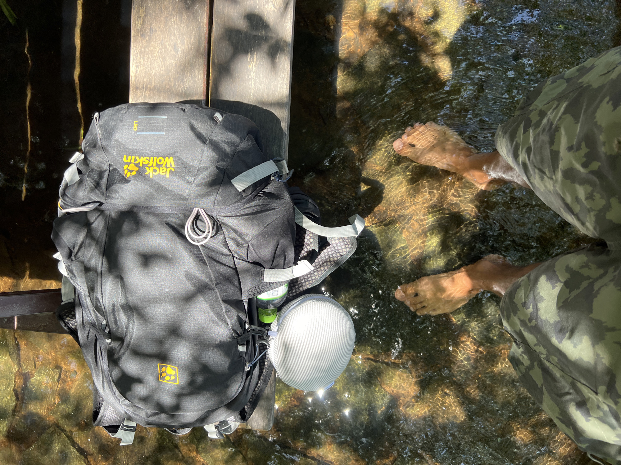 The image displays a first-person, overhead view of a person's lower legs and feet submerged in clear, shallow water, with a backpack resting on a wooden structure to the right. The person's feet are bare, and they are wearing camouflage-patterned shorts.
The water, which occupies the lower two-thirds of the frame, is rippled and reflects sunlight, indicating a bright, sunny outdoor environment with overhead foliage casting dappled shadows. The waterbed is visible, consisting of small stones and earthy tones.
On the wooden structure, a black Jack Wolfskin backpack is placed. The backpack features the brand's yellow paw print logo and text. Attached to the backpack by a blue clip, a light grey, spherical object with horizontal white stripes hangs just above the water. A green plastic bottle is partially visible next to the backpack, near the water. The wooden structure appears to be a bench or a platform alongside the water.
There are no discernible details within the image to confirm the specific location as Nam Cam Ranh, Vietnam. The scene depicts a generic natural outdoor setting, likely a stream or shallow river.