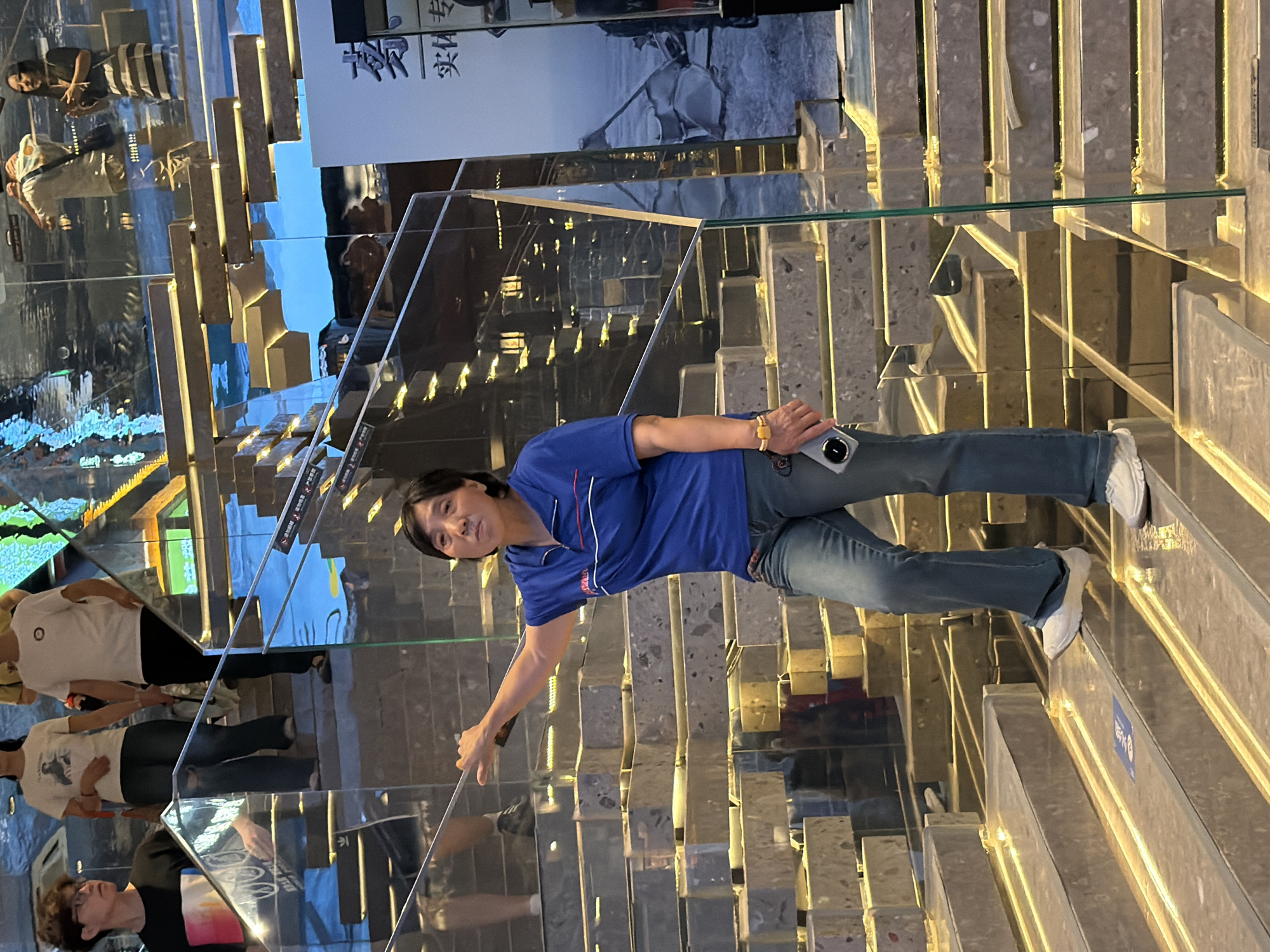 A woman wearing a blue polo shirt and blue jeans stands on a modern, multi-level staircase with reflective surfaces and integrated lighting. She is holding a cell phone in her right hand and her left hand is extended, resting on a glass railing. The woman is looking towards the camera with a neutral expression. Behind her, other people are visible on the stairs, including a man in a black t-shirt and glasses on a higher level. The environment appears to be an indoor public space, possibly a shopping mall or a museum, given the architectural style and the presence of posters on the right side of the image, one of which has Chinese characters. The staircase design features alternating horizontal and vertical elements with embedded warm-toned lights. The reflective surfaces create a visually complex and somewhat distorted environment. The scene is located in the Yuzhong District, China.