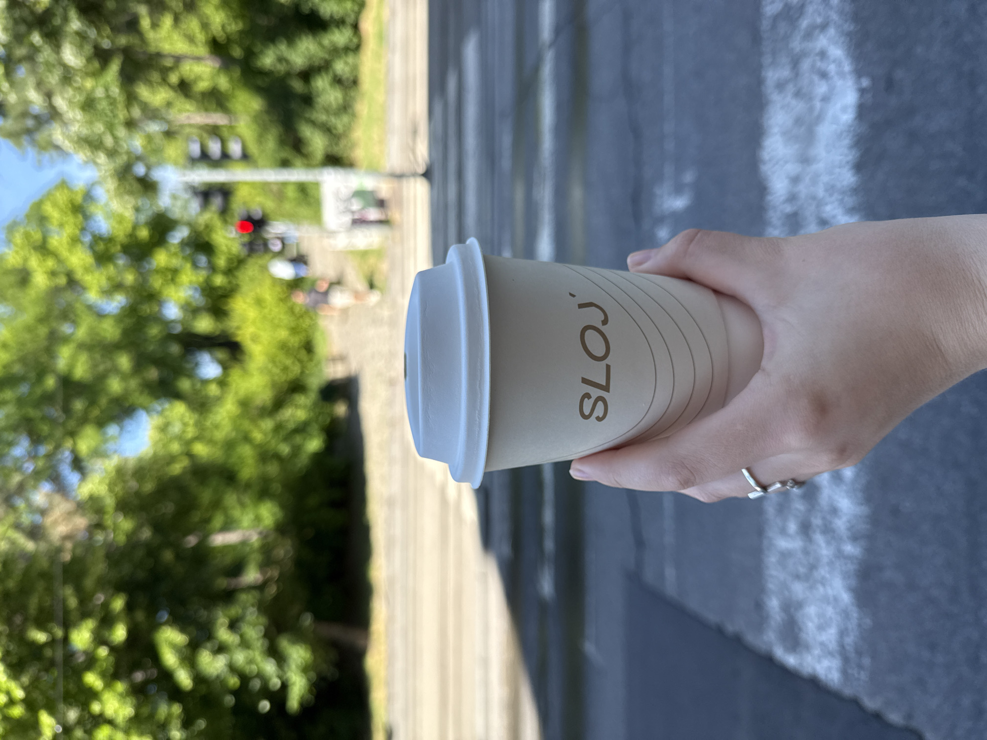 A medium-shot photograph shows a person holding a paper coffee cup with the word "SLOJ," possibly indicating "Layer" in Serbian, which may suggest the name of the coffee shop. The person’s hand is pale-skinned and wears a silver ring on their finger. The cup has a white lid and is held against a blurred background, suggesting depth of field.
The background shows a street with tram tracks. A blurred traffic light is visible, indicating that the light is red. Trees line the street. The picture gives the sense of Belgrade, Serbia, with the combination of tram tracks, urban streetscape, and the Serbian word on the cup. The person is possibly walking or waiting on a street in Belgrade, enjoying their coffee.