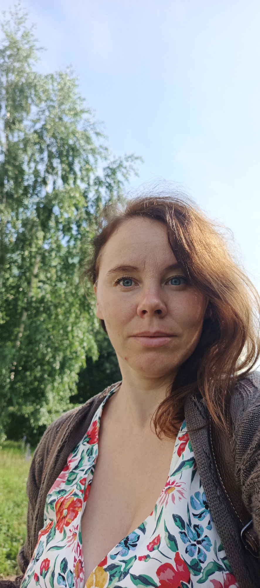A woman with auburn hair and blue eyes is captured in a medium shot outdoors. She is wearing a brown knitted cardigan over a white dress with a colorful floral pattern. The background consists of green trees and a clear blue sky, suggesting a natural, possibly rural or park-like setting. There are no discernible urban structures or landmarks indicating a specific location within the city of Chaikovsky, Russia. The woman is looking towards the camera with a neutral expression. The lighting suggests it is daytime, with sunlight illuminating her face and hair.