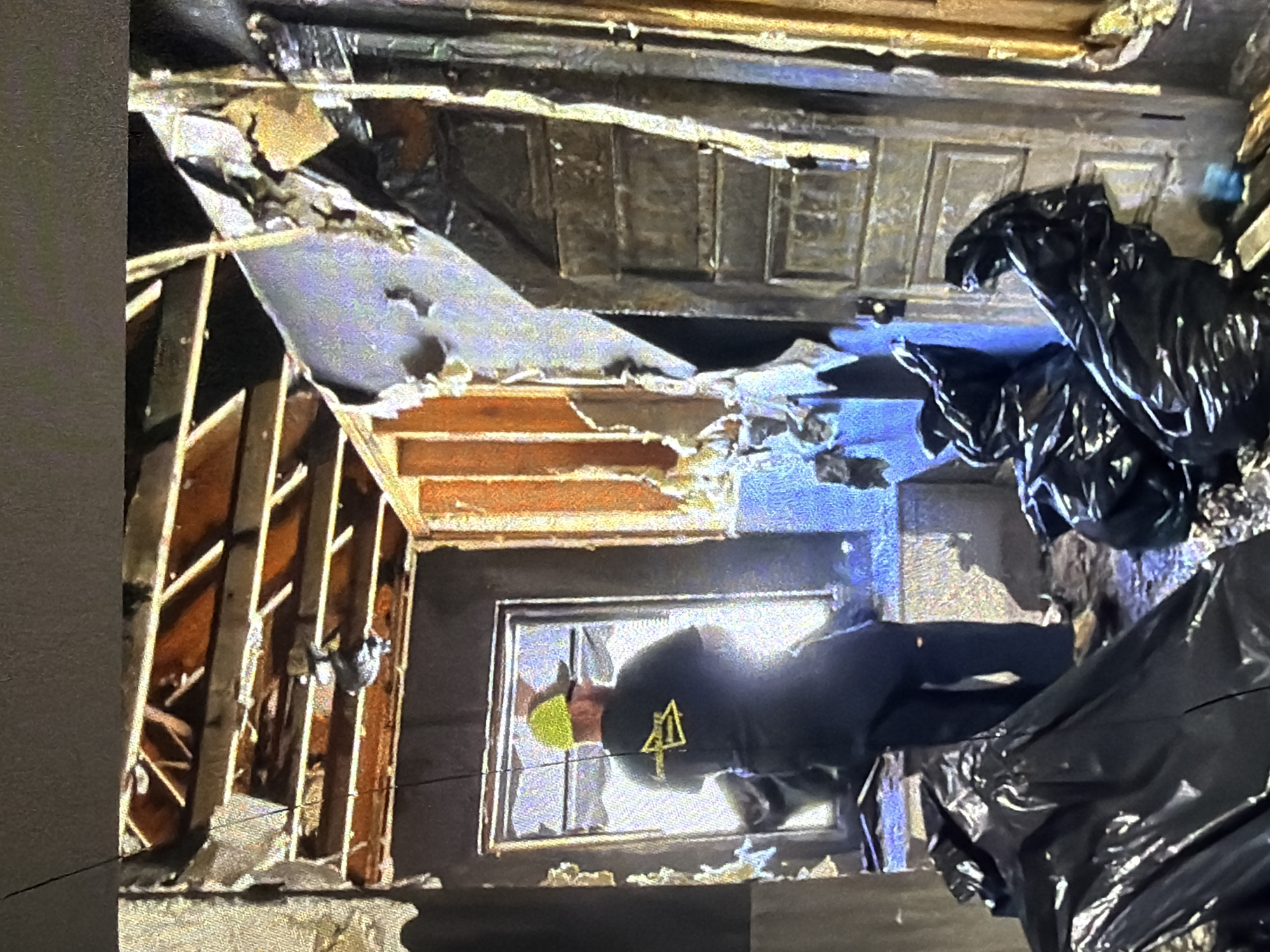 In Bukuru, Nigeria, a person wearing a yellow hard hat and a black t-shirt with a yellow logo is standing in a severely damaged room. The ceiling joists are exposed, and sections of the walls and ceiling are missing or heavily damaged, with debris visible. Black trash bags are scattered on the floor. A door frame and a window with a view of bright light are also present in the scene.