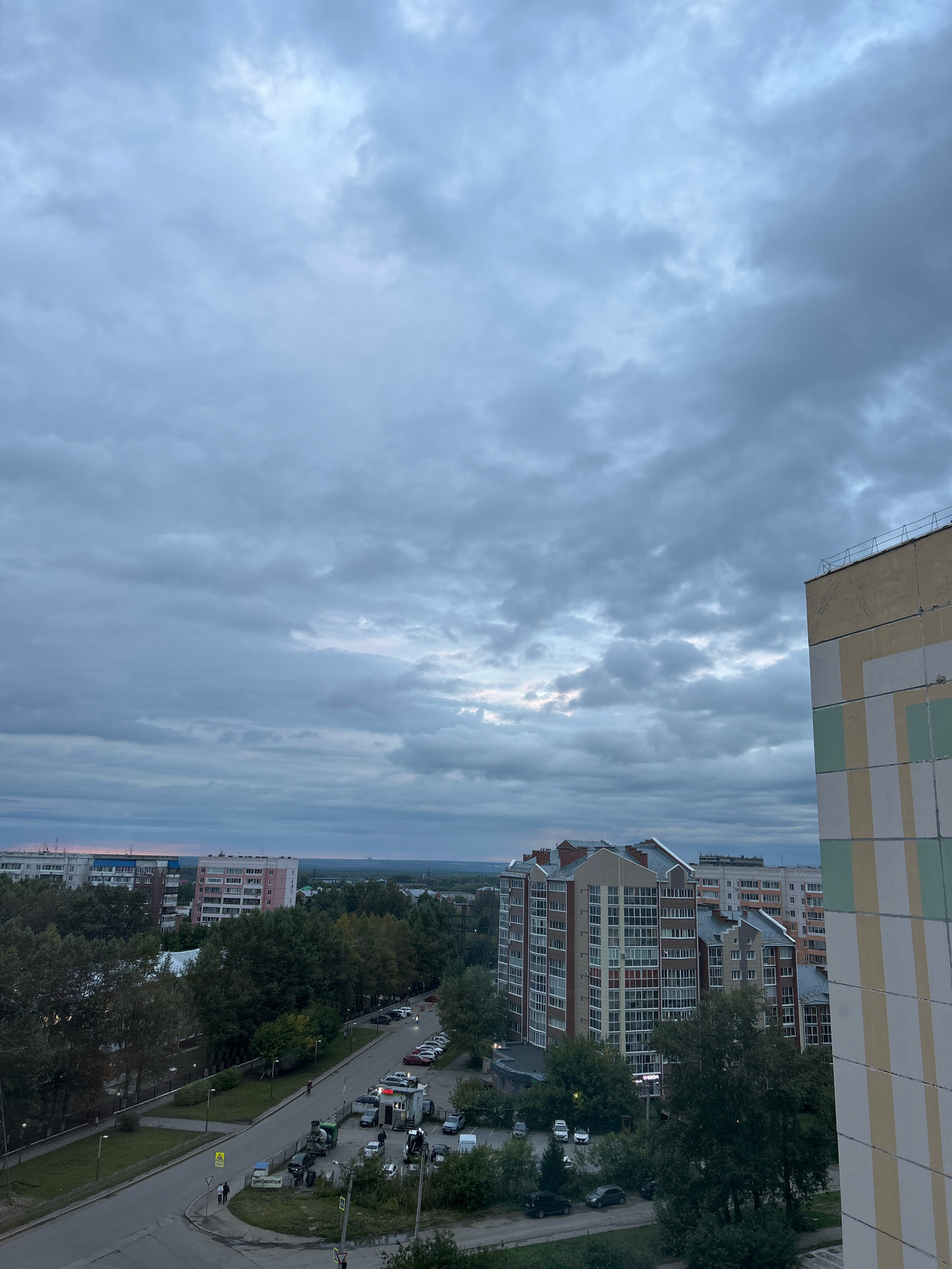 The image shows an overcast sky above a cityscape. In the foreground, the side of a building with a geometric pattern of yellow, green, and white panels is visible on the right. To the left of this building, a street curves through a neighborhood with several multi-story apartment buildings, trees, and parked cars. Further in the distance, more apartment buildings and a line of trees can be seen against the horizon. The sky is filled with dense, grey clouds, with a faint hint of pinkish light visible near the horizon in the distance, suggesting either dawn or dusk. The overall impression is of a typical urban scene under a cloudy sky. Based on the architectural style of the buildings and the general appearance, this scene is consistent with urban areas in Tomsk, Russia.