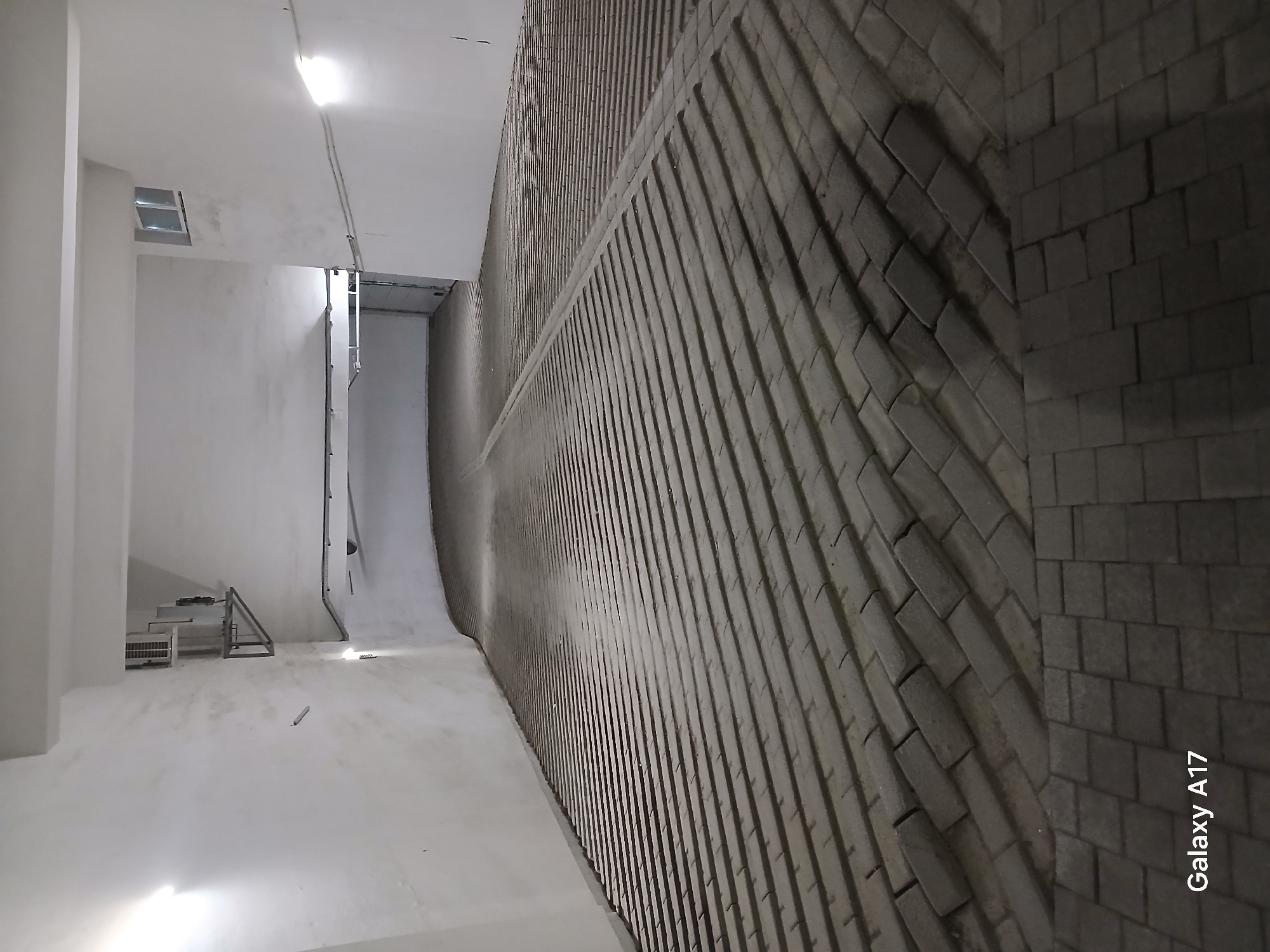 The image depicts an indoor space, possibly a building interior, with an unusual architectural design. The floor and walls are primarily white, contrasting with a large curved wall constructed of textured, gray brick. The brickwork transitions from textured to standard blocks. The curved wall suggests a bend in the structure. In the background, near the left, a window-like opening is visible, along with some metallic structures and pipes. The overall environment appears clean and utilitarian. The watermark "Galaxy A17" in the lower right corner indicates the image was taken with a Samsung Galaxy A17 smartphone. There are no people visible in the scene. The location is Yekaterinburg, Russia.