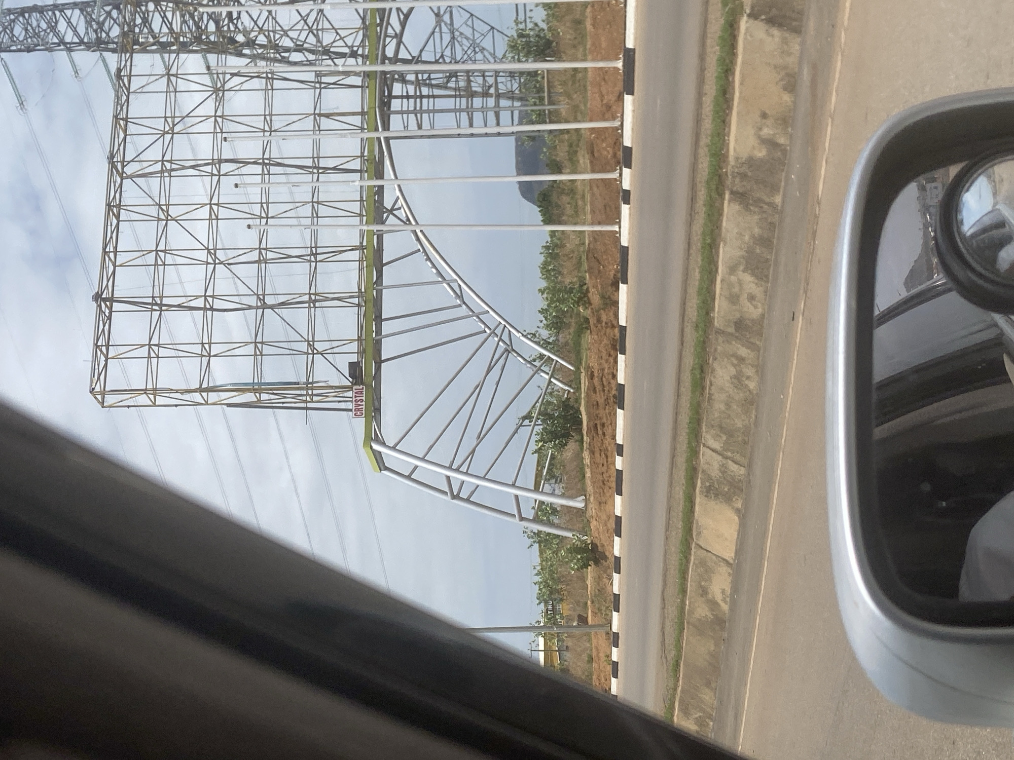 The image shows a view from inside a vehicle, with the side mirror partially visible on the right. A large metal structure, likely a billboard support, stands alongside a road. The billboard is labeled "CRYSTAL" and is located in Abuja, Nigeria. The roadside has vegetation and a curb with black and white markings. Power lines are also visible in the background. The sky is overcast. The road appears to be paved and in good condition.