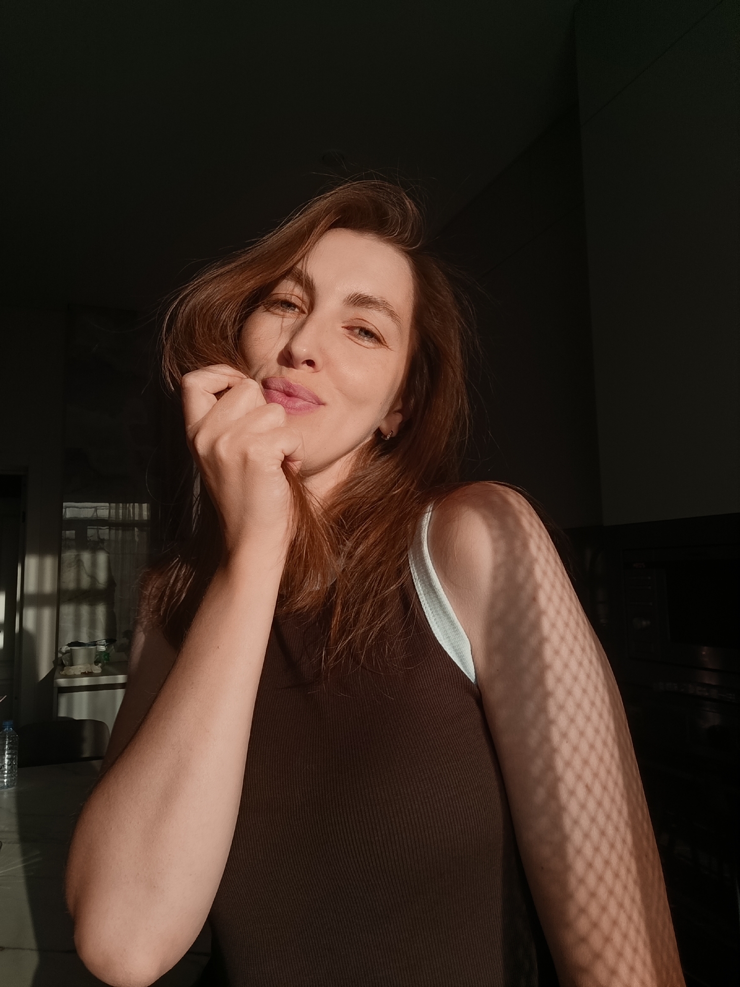 This is a medium shot, portrait-style photograph of a young woman indoors, bathed in warm, directional sunlight that creates dramatic shadows. She has reddish-brown hair that is slightly tousled and cascades over her shoulders. Her right hand is raised, gently touching her lips and chin, with her fingers partially obscuring her mouth. She is looking towards the camera with a soft, slightly playful expression, her eyes are open and her lips are slightly parted.
She is wearing a brown, ribbed tank top with a lighter colored strap visible on her left shoulder. The sunlight casts a distinct criss-cross shadow pattern on her right arm and shoulder, suggesting the presence of blinds or a lattice structure outside a window. The background is dark and out of focus, with some subtle hints of interior space, possibly a room with furniture. The strong sunlight, casting long shadows and illuminating her face, suggests it is either early morning or late afternoon. The overall mood is intimate and contemplative.
There is no visible text in the image. The location context of Chaikovsky, Russia, is noted but not visually indicated within the image itself.