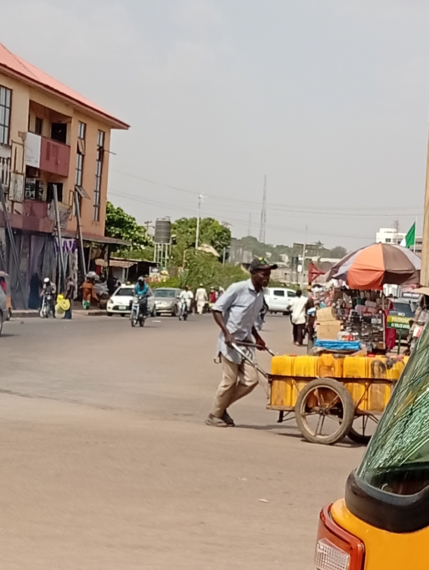 The image shows a street scene in Maiburiji, Nigeria. A man in a light blue shirt and a cap is pulling a two-wheeled cart laden with yellow containers, likely water jugs, across an intersection. The environment appears to be a busy commercial area with buildings, vehicles, pedestrians, and vendors. There are motorcycles and cars in the background. The sky is overcast.