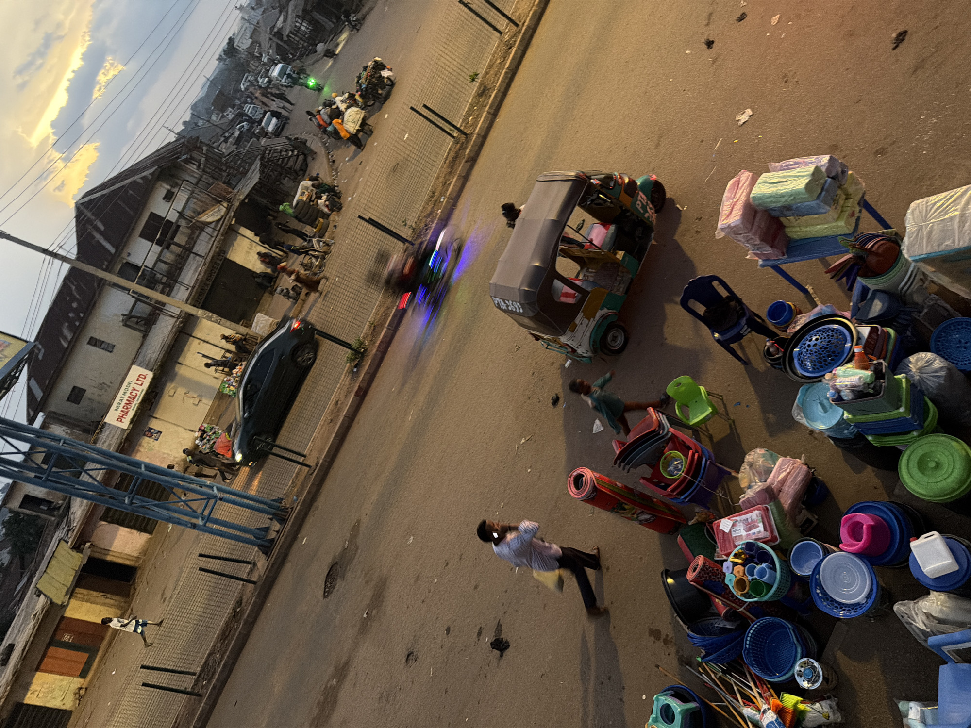 The image captures a street scene in Bukuru, Nigeria, during what appears to be late afternoon or early evening, indicated by the warm, low-angle sunlight casting long shadows. The foreground is dominated by a collection of assorted plastic household items, including buckets, basins, bowls, and chairs, piled on the ground and on a makeshift cart. A man in a patterned shirt and dark pants is walking on the street, holding a light-colored bag and appearing to be on his phone. To the right, a child in a green top runs across the street.
In the middle ground, a three-wheeled auto-rickshaw, known locally as a "Keke Napep," is parked on the street. Several other vehicles, including cars and motorcycles, are visible further down the road, suggesting moderate traffic. The buildings lining the street are a mix of commercial establishments and residential structures, some appearing weathered with corrugated iron roofs. A prominent sign reads "NERAT MODEL PHARMACY LTD."
The street itself is paved and shows signs of wear, with some cracks and debris. A metal fence runs along the sidewalk in some sections. The background reveals a dense urban landscape with more buildings stretching into the distance, and the sky is filled with dramatic, illuminated clouds. The overall impression is one of a bustling, everyday commercial activity in a Nigerian urban setting.