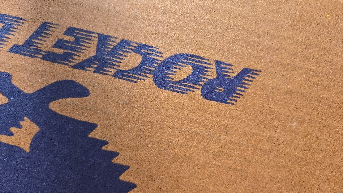 A close-up, vertically oriented image displays a textured, light brown substrate, consistent with cardboard or a fibrous mat. Dark blue graphic elements are printed directly onto this surface. The word "ROCKET" is partially visible, oriented diagonally upwards from the lower right to the upper left. The letters 'R', 'O', 'C', 'K', 'E', and a portion of 'T' are discernible. Each letter features a distinct fringed or serrated pattern along its lower contour. To the upper left of the visible text, a partial dark blue graphic with a jagged, irregular outline is present, resembling a stylized avian or mythical creature. No individuals, animals, or observable actions are depicted. The environment is an unspecified setting within the city of Unknown, Unknown.