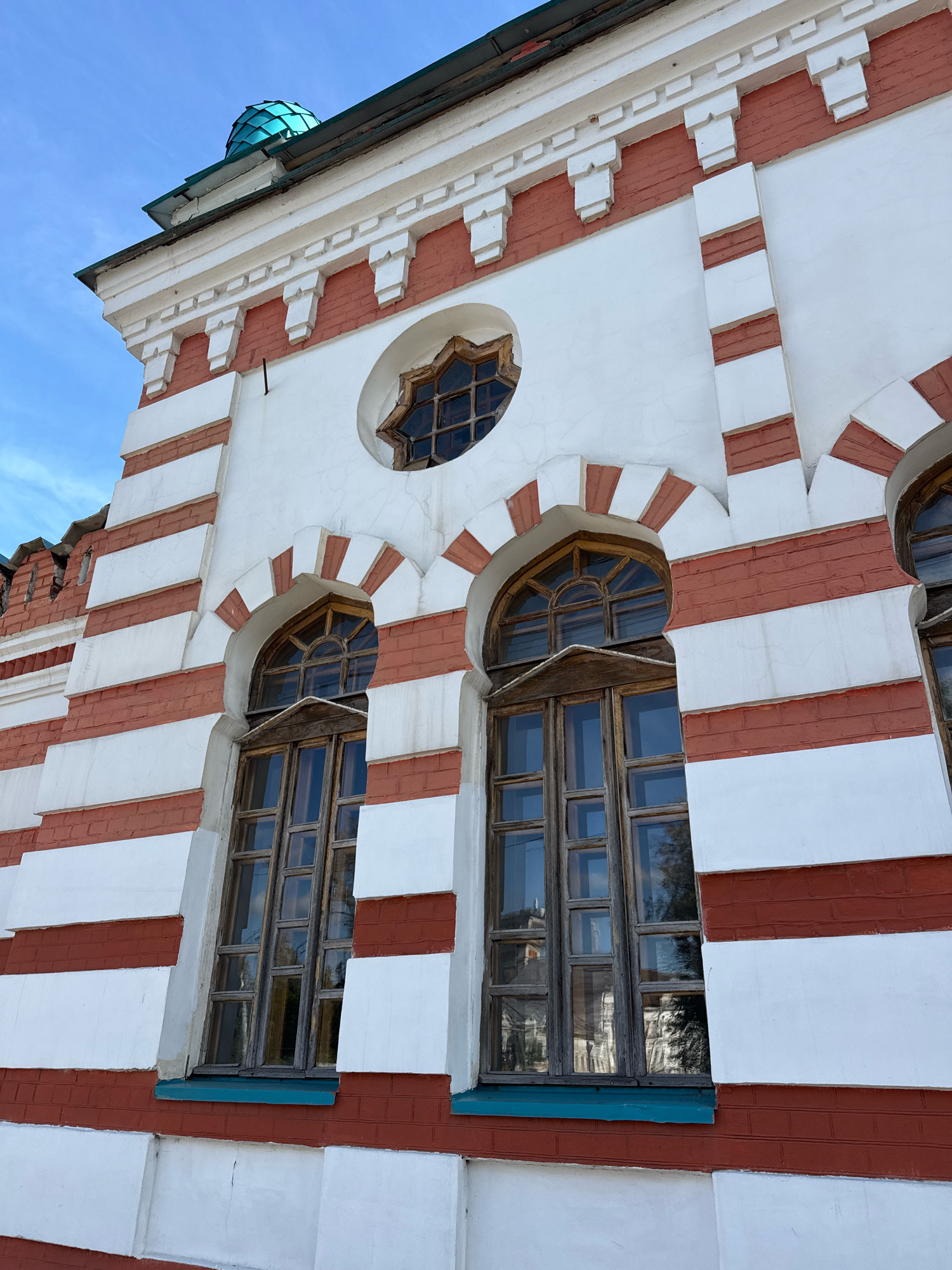 The image shows a close-up, low-angle view of the facade of a building with red and white horizontal stripes and white decorative elements. Two large arched windows with wooden frames and multiple panes are visible. Above the windows, there is a small circular window with a star-shaped frame. The roofline features a decorative cornice and a small, teal-colored dome. The building appears to be made of brick, with sections painted white. The sky is blue with a few wispy clouds. The architectural style suggests it could be a historical or religious building. This building is located at the Church of the Nativity of John the Baptist in Tver, Russia.