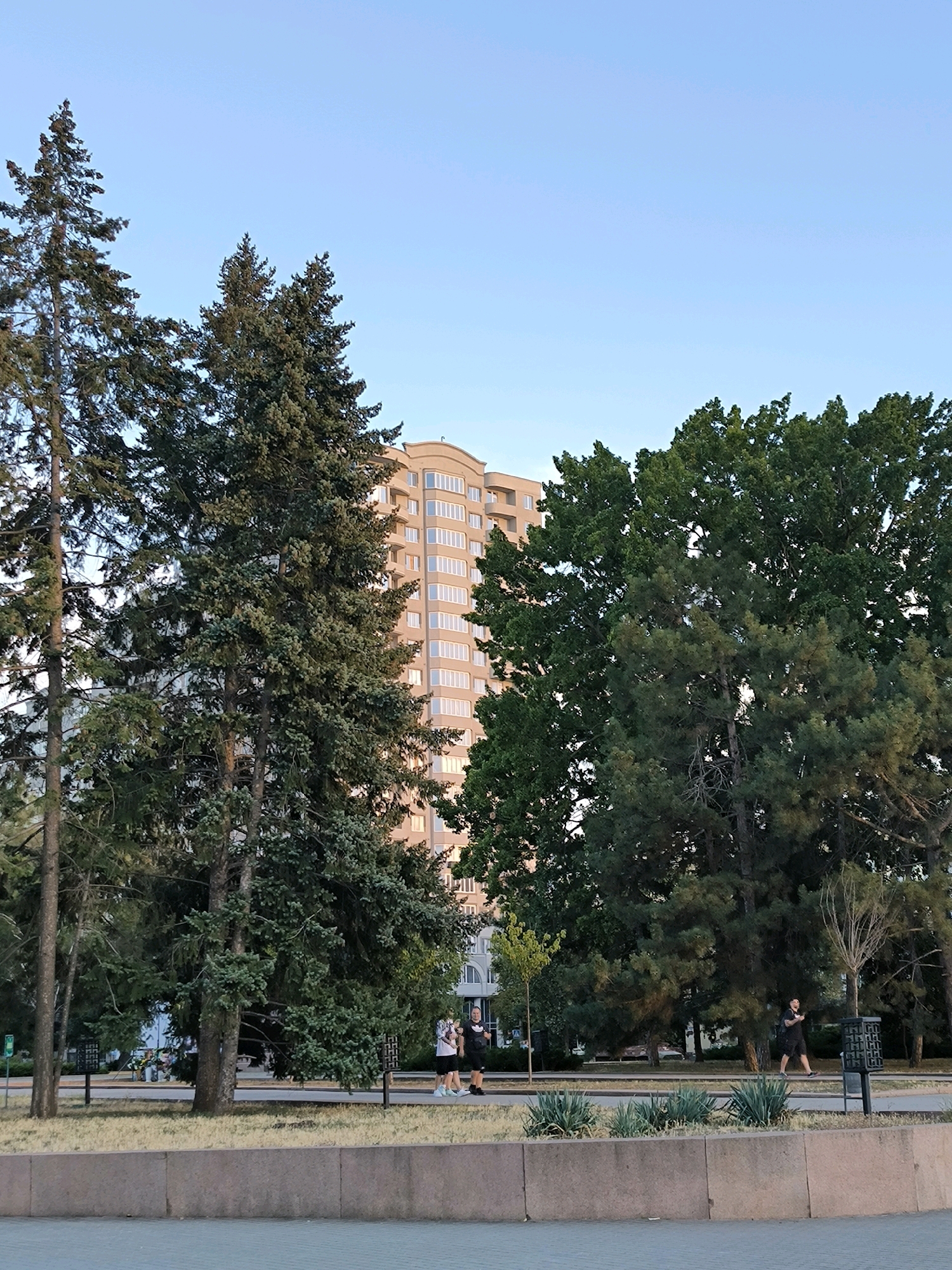 The image shows a park area with mature trees and a modern apartment building in the background. Two individuals are walking on a paved path. One person is wearing a black t-shirt and shorts, while the other is wearing a white t-shirt and dark shorts. The apartment building appears to be a multi-story structure with numerous windows. The park features a low concrete border in the foreground and grassy areas with scattered bushes. The sky is clear and blue, suggesting daytime. The presence of tall trees and the urban architecture points to a developed city environment. This scene is consistent with a public park in Mykolaiv, Ukraine.