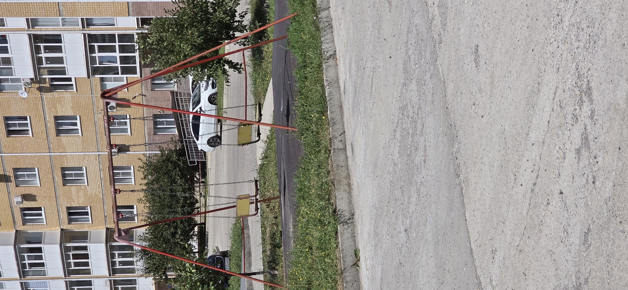 A playground swing set with two yellow seats is in the foreground. The swing set has a red metal frame. Behind the swing set is a sidewalk, grass, trees, and a building. The building is made of brick and has balconies and windows. A white car is parked in front of the building. A dark-colored car is parked to the left of the swing set. The image appears to be taken on a sunny day. This scene is located in Inozemtsevo, Russia.