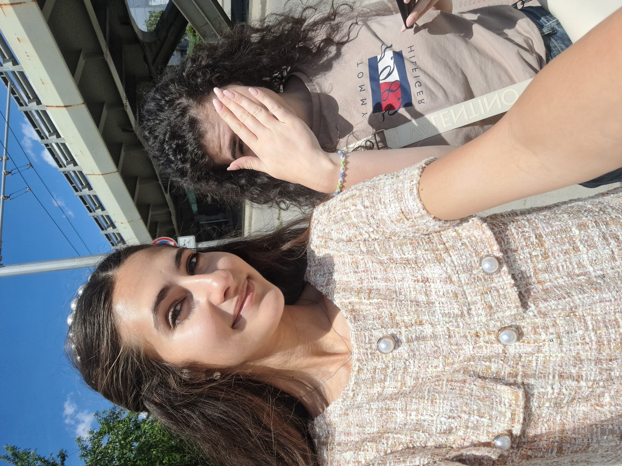 Two young women are captured in a selfie-style photograph taken outdoors. One woman is in the foreground with long, wavy brown hair adorned with a pearl-studded headband and wearing a tweed-like jacket with pearl buttons. She is smiling slightly and looking directly at the camera. In the background, the second woman, with dark curly hair, covers part of her face with her hand. She wears a beige Tommy Hilfiger shirt and is holding a smartphone in her other hand. A colorful beaded bracelet is visible on her wrist.
The scene is under a bridge structure, evident from the steel beams and supports visible at the top of the frame. A blue sky can be seen between the beams. A traffic sign is partially visible. Green foliage from trees is at the bottom left. The location is identified as the city of Sofia, Bulgaria, based on traffic signs and street markings.