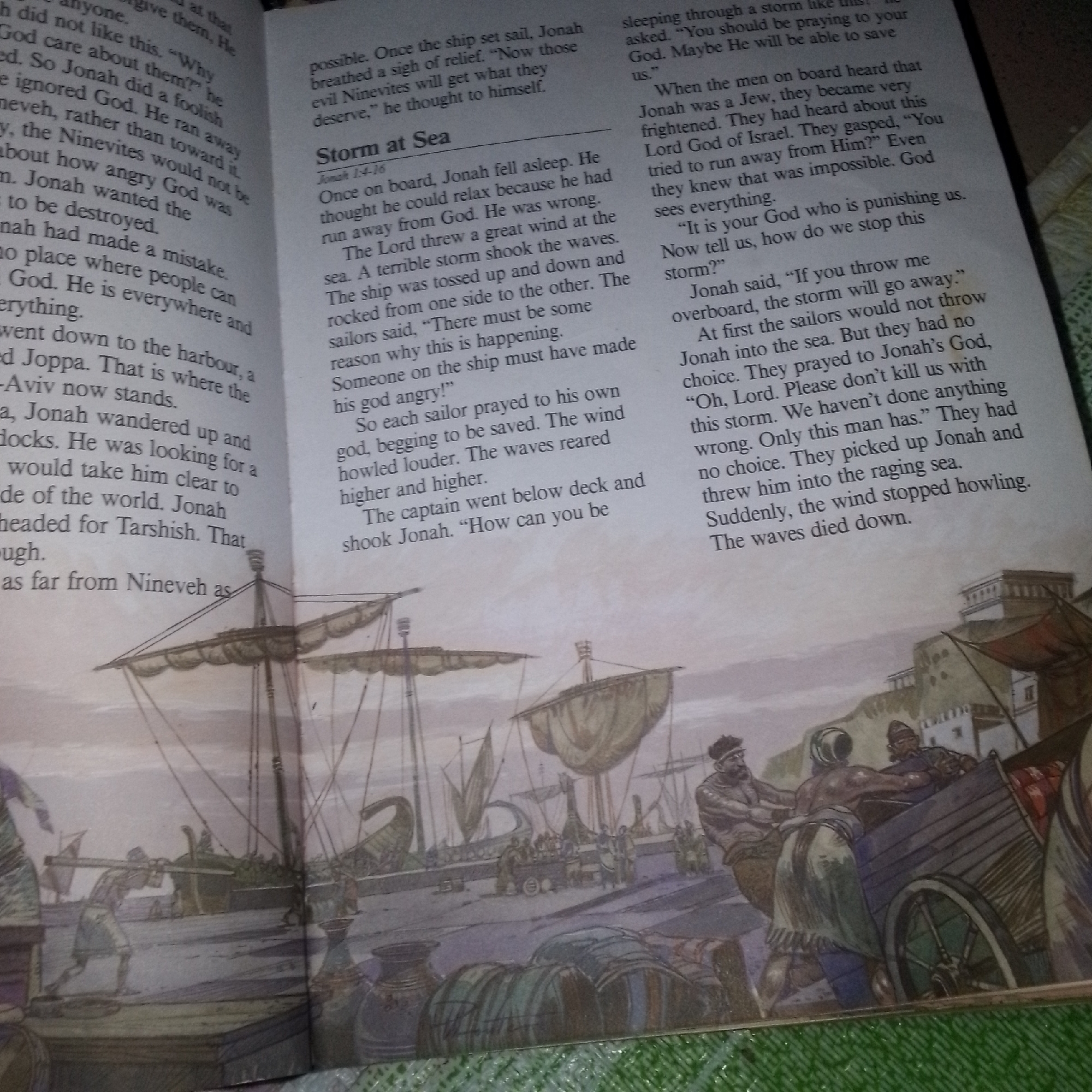 The image depicts an illustration from a book, showing a biblical scene of a ship and sailors. On the left side, text discusses Jonah and his journey. The right side of the image shows a caption "Storm at Sea" with related text. The illustration features several ships with sails, docked at a port with buildings in the background. People are visible on the dock, some appearing to be loading cargo onto a cart. The overall scene suggests a bustling port during a time of storm, as alluded to by the text. The specific location is not identifiable from the image itself, but the text refers to Joppa.