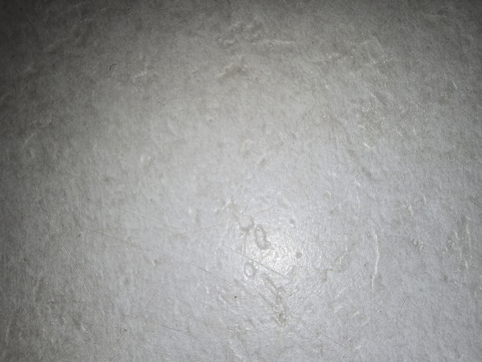 The image shows a close-up, top-down view of a textured, light gray surface, possibly a floor or countertop. The surface appears to be somewhat worn, with visible scratches and slight imperfections. There are areas where the surface is brighter, suggesting it might be slightly wet or reflective. The overall lighting creates a gradient effect, with the lower portion of the image being brighter and the upper portion becoming darker. No people, specific objects, or discernible environmental context are visible. The image is devoid of any identifiable landmarks or features that would place it in Ulan-Ude, Russia, or any other specific location.