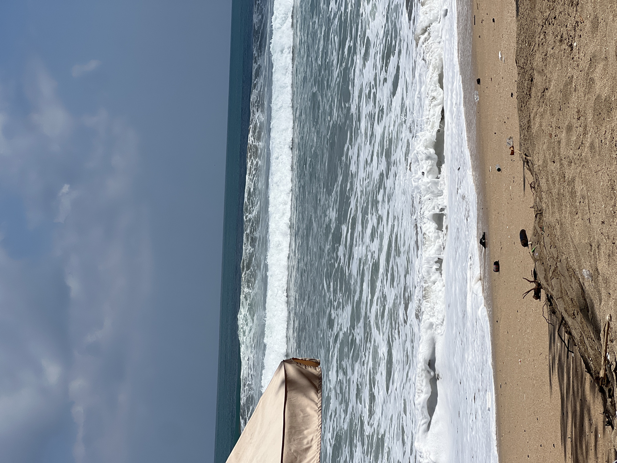 The image captures a segment of the beach in Kuta, Indonesia, characterized by its sandy shoreline and the rhythmic movement of waves. In the foreground, the beach is a mixture of sand and some debris, including pieces of driftwood and scattered plastic waste. A beige beach umbrella partially obscures the left side of the frame, casting a shadow on the sand. The ocean is the dominant feature, with multiple rows of white-capped waves rolling towards the shore. The water transitions from a lighter turquoise in the foreground to a deeper blue towards the horizon, where it meets a hazy sky with scattered clouds. The overall atmosphere is that of a slightly overcast day at a tropical beach.