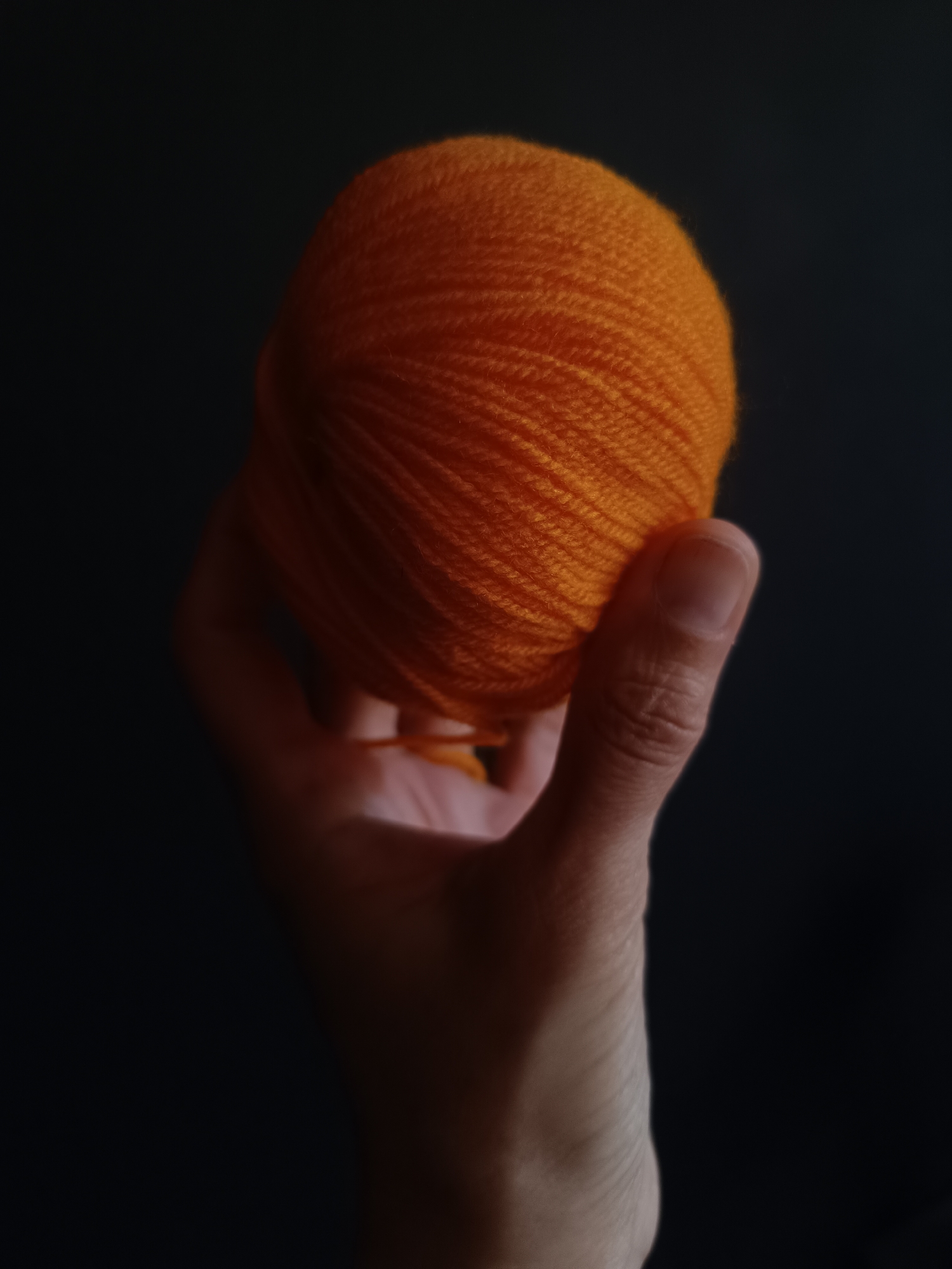 A human hand, likely an adult's, is prominently featured holding a vibrant ball of orange yarn against a uniformly dark, possibly black, background. The scene is illuminated from the front, highlighting the textured surface of the yarn and the contours of the hand, which appears to be reaching towards the viewer. The bright orange of the yarn creates a strong contrast with the deep, shadowed background, making it the central focus of the image.
The yarn itself is tightly wound, showcasing its soft, slightly fuzzy texture and numerous individual strands. Details of the hand are visible, including skin texture and the fingernail of the thumb, suggesting a clean and well-kept hand. No specific activity beyond holding the yarn is depicted, giving the image a still-life quality. The lighting suggests an indoor setting or a controlled environment, as no external light sources or weather conditions are discernible. There is no visible text within the image. The minimalist background emphasizes the simplicity and material quality of the yarn and the human element.