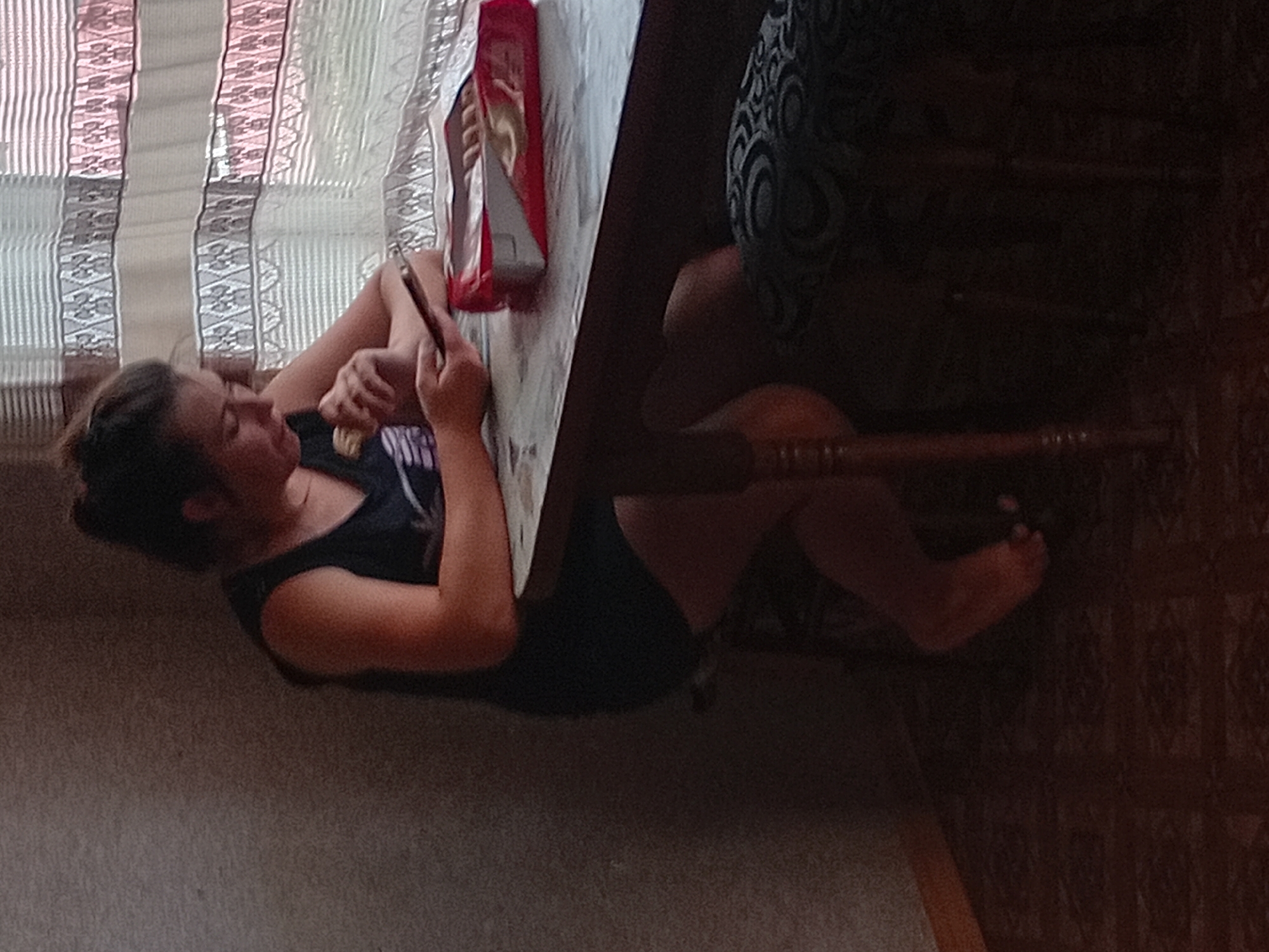This media file depicts a young person, likely female, sitting on the floor with their legs tucked up, leaning against a wooden table. The person is looking down at their phone, which they are holding with both hands. They are wearing a dark tank top. To the left of the person, a window is visible, covered by vertical blinds with a decorative pattern. On the table, a red and white package, possibly of snacks, is present. The scene appears to be indoors, with the floor covered in carpet and a patterned chair or cushion visible in the background. The lighting suggests it is daytime. The overall impression is one of casual relaxation and engagement with a mobile device. The scene is set in Verkhniachka, Ukraine.