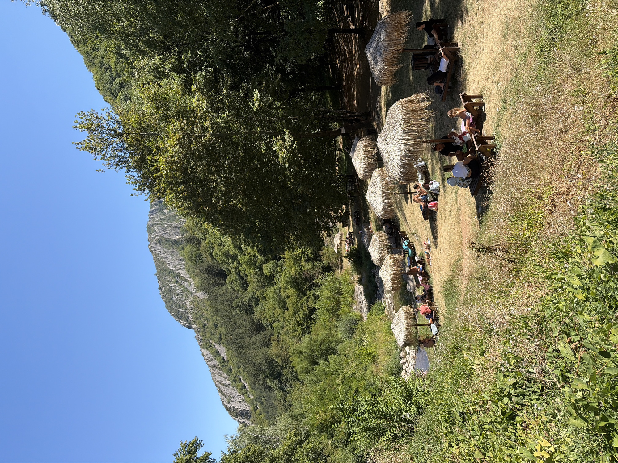 The image depicts a scenic outdoor recreational area at a location near Zgorigrad, Bulgaria. A clear blue sky dominates the upper portion of the frame, above a rugged, tree-covered mountain range. In the foreground and middle ground, a grassy area is dotted with several thatched umbrellas providing shade for lounge chairs. Several individuals are relaxing on these chairs, some in groups, others individually. A small stream or river flows through the scene, with rocky banks visible. Lush green vegetation, including trees and shrubs, surrounds the recreational area, creating a natural and tranquil ambiance. The overall scene suggests a peaceful day at a popular outdoor spot.