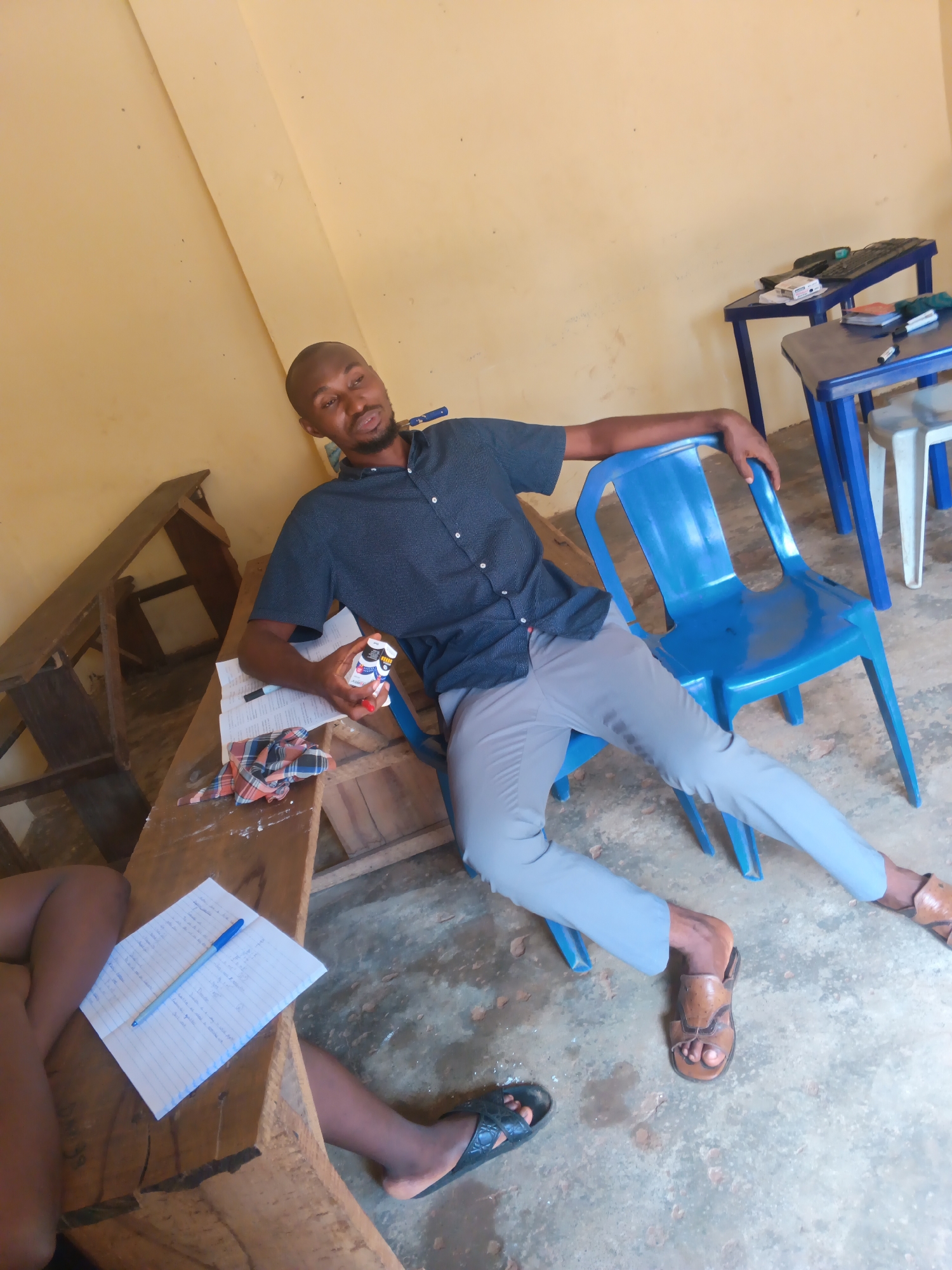 The image depicts a man sitting on a blue plastic chair in what appears to be a classroom or study area in Onitsha, Nigeria. He is wearing a dark patterned short-sleeved shirt and light grey trousers, with brown leather sandals on his feet. He is holding a small white and red bottle in his right hand. In front of him, on a wooden desk, is a notebook with a blue pen, some papers, and a checkered piece of cloth. Other wooden desks and chairs are visible in the background, as well as two blue tables with what seem to be computer equipment. The walls are painted a light yellow color, and the floor is concrete with some debris. Another person's legs are visible in the lower left corner of the frame, wearing black sandals and resting on a wooden surface. The lighting suggests an indoor environment, possibly during daylight hours.
