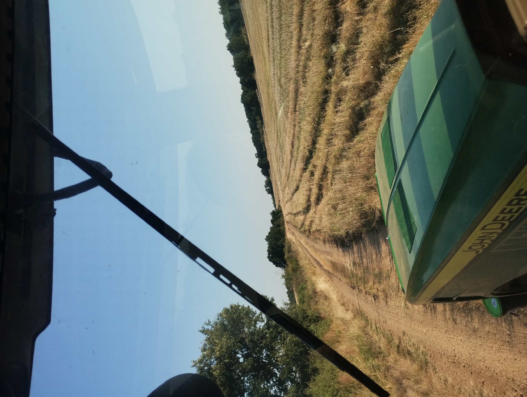 The image depicts a view from the cab of a green John Deere tractor, traveling on a dirt road through a harvested field. The tractor's hood is visible in the foreground, with the "JOHN DEERE" logo clearly displayed. The field, a golden-brown color indicating dry crops, has visible tire tracks from harvesting equipment. A treeline borders the field in the distance, with a clear blue sky overhead. A windshield wiper is present in the upper portion of the image. The scene appears to be in a rural setting, likely the city of Kubadin, Bulgaria, given the context. There are no people or animals visible in the image.