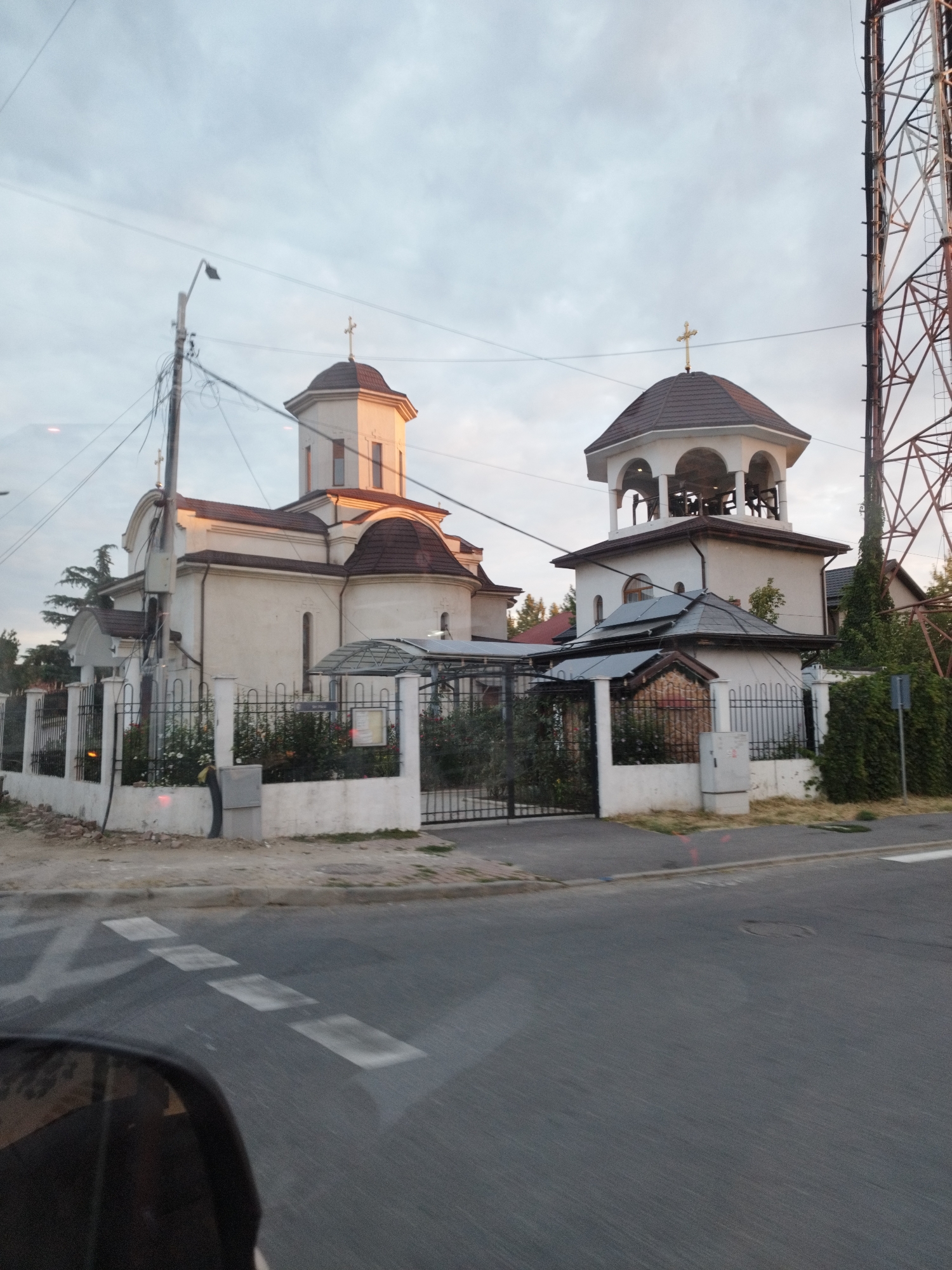 The image shows a church with a tall bell tower, located in Chitila, Romania. The church is painted white with a brown roof and has several windows and crosses. The bell tower is octagonal and has an open structure with bells visible inside. A fence surrounds the church property, with a gate in the foreground. There are trees and bushes around the church, and a street with a pedestrian crossing in front of it. A large telecommunications tower is visible in the background on the right side of the image. The sky is cloudy and the light suggests it might be late afternoon or early morning. The image appears to be taken from a moving vehicle, as indicated by the slight motion blur and the reflection in what appears to be a car mirror in the lower left corner.