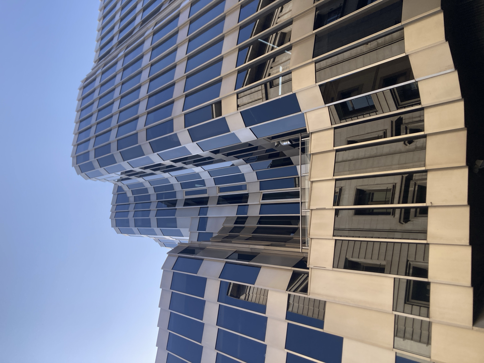 The image shows a modern building with a curved facade made of glass and metal panels. The building is angled upwards, with a clear blue sky in the background. The glass panels reflect the sky and the surrounding buildings, creating a sense of depth and dynamism. In the lower part of the image, the reflection of an older, more traditional building with windows and architectural details is visible in the glass panels. The architectural style of the building, particularly the curved glass facade and the metallic accents, is characteristic of contemporary office buildings found in business districts of major cities.
Without further information or context, pinpointing the exact location of this building in Barcelona, Spain is not possible based solely on the visual evidence. However, such an architectural style is common in the newer commercial areas of Barcelona, like the area around Diagonal Mar or the business district of La Nova Esquerra de l'Eixample, which feature a mix of modern and historic architecture.
There are no people, animals, or specific actions visible in the image. The focus is entirely on the architectural design of the building and its interaction with light and reflection. The environment depicted is urban, with the presence of other buildings suggested by the reflections.