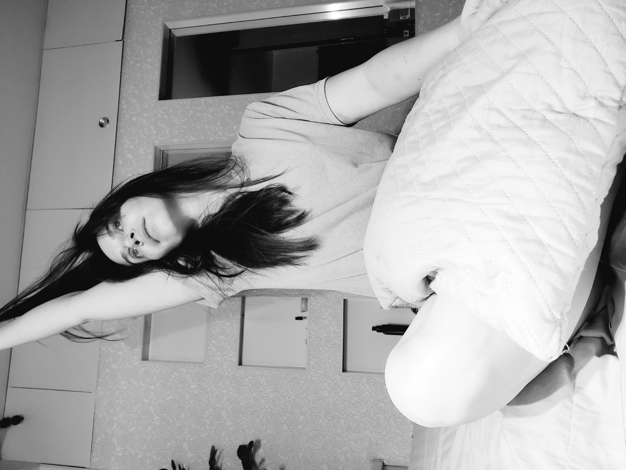 A grayscale image shows a woman lying on a bed with her head tilted towards the left and her right arm extended upwards. She is wearing a light-colored shirt. A white quilted object covers the right side of the image. The background includes a floral-patterned wall with two square picture frames, and a wardrobe with a small window on top. There is no evidence to support the claim that the location is in the city of Chaikovsky, Russia.