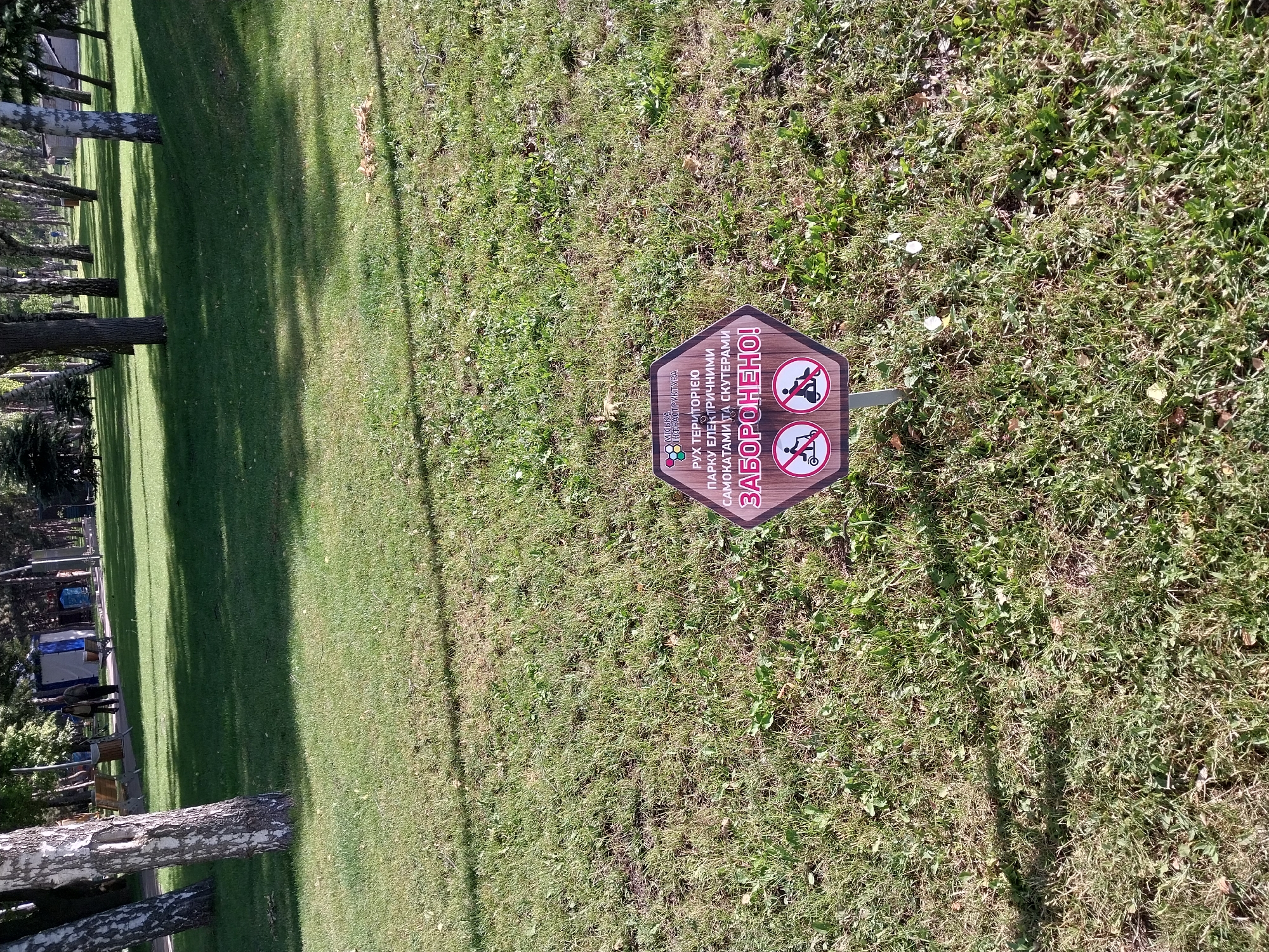 A hexagonal sign is planted in the grass of a park. The sign is brown and has text and icons on it. The text reads "МІСЬКА ІНФРАСТРУКТУРА" (City Infrastructure) at the top, followed by "РУХ ТЕРИТОРІЄЮ ПАРКУ ЕЛЕКТРИЧНИМИ САМОКАТАМИ ТА СКУТЕРАМИ ЗАБОРОНЕНО!" (Movement of electric scooters and scooters through the park is PROHIBITED!). Below the text are two circular icons, both with a red circle and a diagonal red line through them. The left icon depicts a person on a kick scooter, and the right icon depicts a person on a motorized scooter or moped. The sign is located in a park with trees and a grassy lawn. In the background, there are more trees, park benches, and some structures. The image appears to have been taken during the daytime.