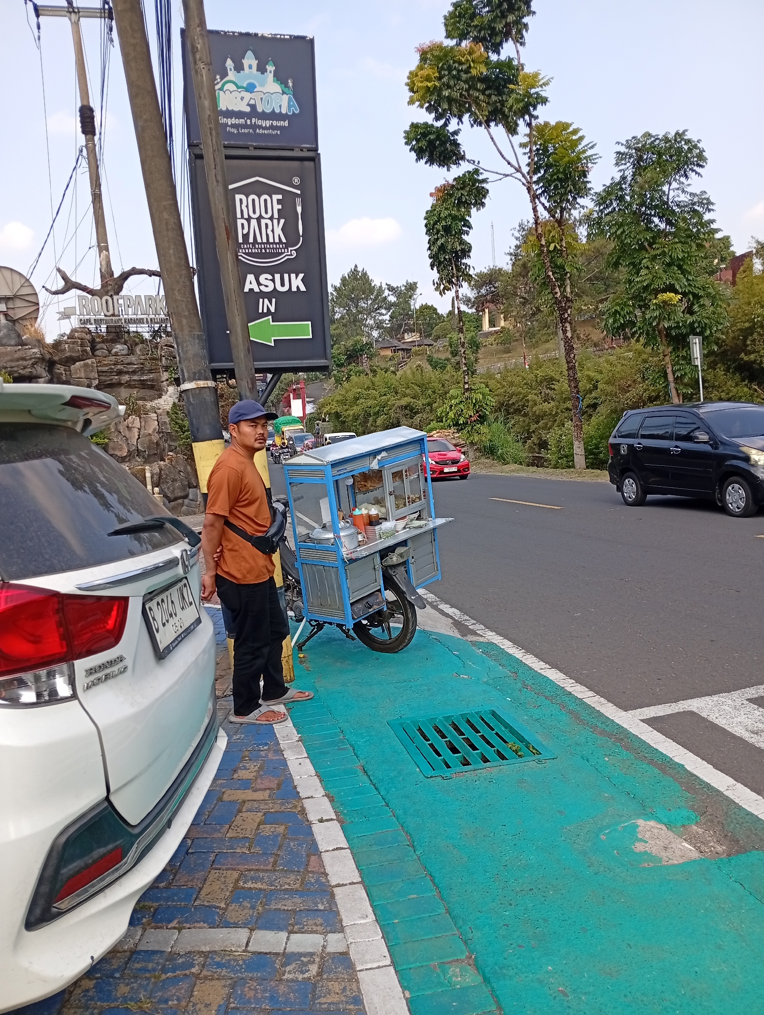 In Cimacan, Indonesia, a man stands beside a white car and a food cart on a street. The food cart, which appears to be on a motorcycle base, is blue and white and stocked with various items. The man is wearing an orange t-shirt, black pants, a blue cap, and a black fanny pack. He is looking towards the right of the frame. The street has a vibrant green painted lane and a brick pedestrian walkway with blue and grey tiles. Traffic signs are visible in the background, including one for "ROOF PARK" with an arrow pointing left. A black car is driving on the road to the right. The environment is characterized by trees, some of which are tall and leafy, and a clear sky. The overall scene depicts a roadside setting with everyday activities.