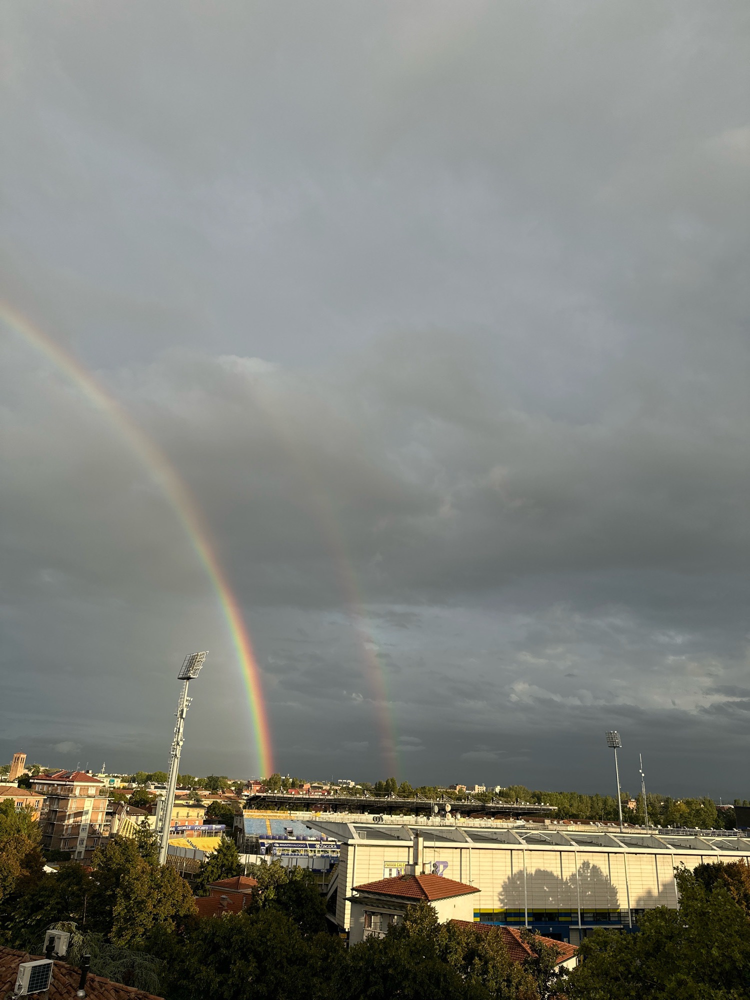 The image captures a dramatic sky over the city of Parma, Italy, featuring a prominent double rainbow against a backdrop of heavy, grey clouds. In the foreground, the rooftops of buildings and lush green trees are visible, indicating an urban setting. Dominating the middle ground is a stadium, likely the Stadio Ennio Tardini, identifiable by its tiered seating in yellow and blue and a long white facade with numerous windows. A tall floodlight tower stands near the stadium, silhouetted against the stormy sky. In the distance, other city buildings and a church tower can be seen. The presence of trees suggests it is likely autumn. No people are visible in the frame. The overall scene conveys a sense of a recent or impending storm, with the natural phenomenon of the double rainbow offering a striking visual contrast to the overcast conditions.