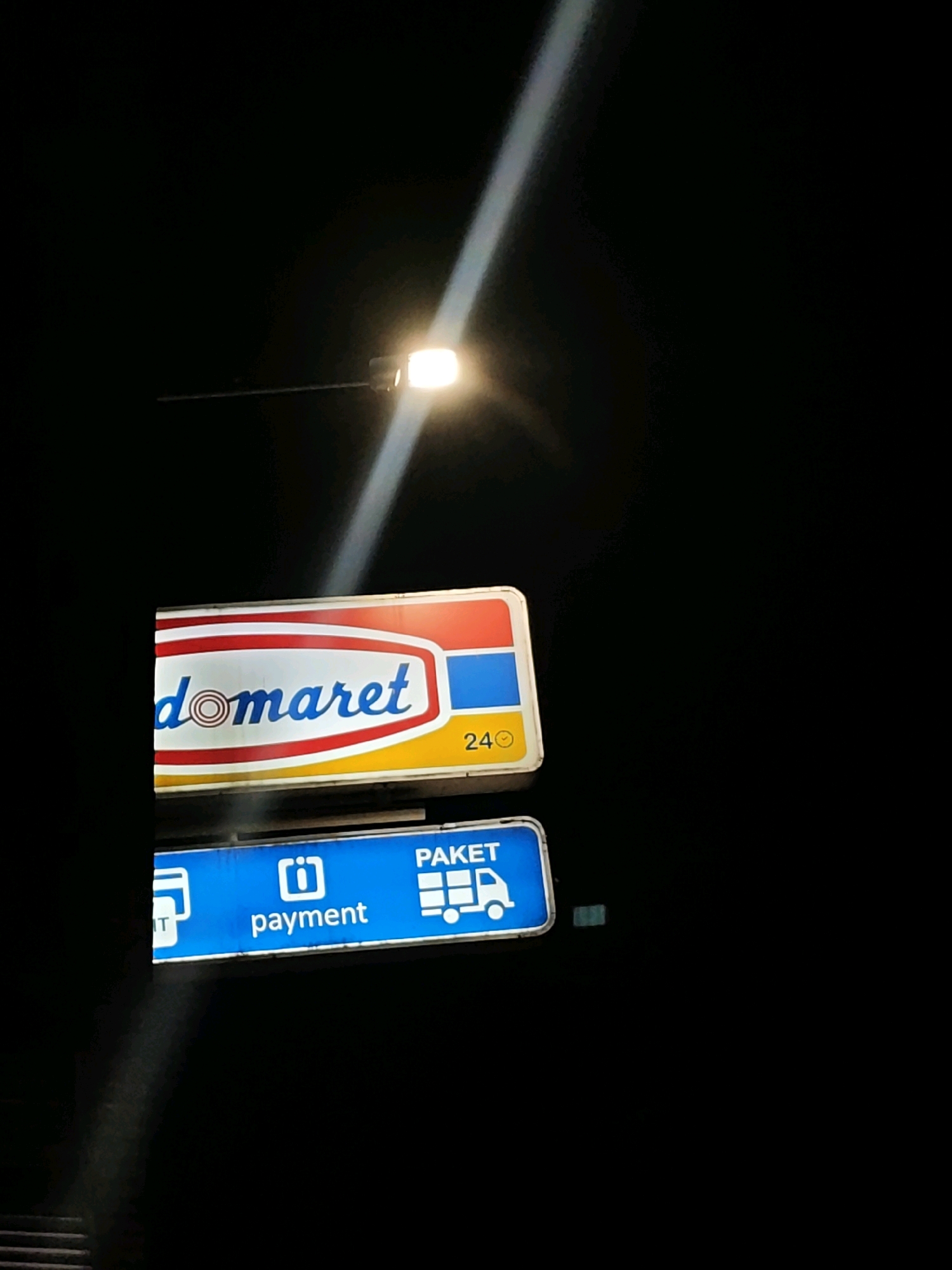 The image captures a dimly lit scene at night, featuring two illuminated signs for a "domaret" convenience store in Gumuk Mas, Indonesia. The top sign, with a red, white, blue, and yellow color scheme, displays the "domaret" logo prominently in blue lettering with a concentric circle design at its center. To the right of the logo, the number "24" is visible, suggesting the store operates 24 hours. Below this, a second, blue sign features white text and icons. On the left, a credit card icon is partially visible, followed by the word "payment" with an information icon. To the right of "payment," the word "PAKET" is displayed above a graphic of a delivery truck, indicating package services. The signs are lit by an overhead street lamp, which casts a bright beam of light diagonally across the dark sky. The surrounding environment is dark, with no other discernible features visible.