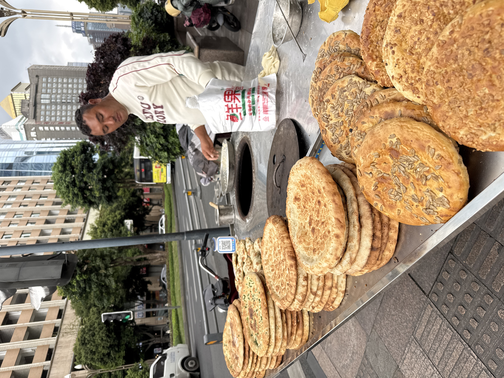 A street vendor in the Qingyang District of Qingyang, China, is selling naan. The vendor, an adult male, is wearing a white sweatshirt with maroon text. He is reaching into a white plastic bag labeled with Chinese characters. A tandoor oven is on the vendor's cart next to the bag. The naan is arranged in stacks on a stainless steel countertop. Some naan is topped with seeds or herbs. The cart is parked on a sidewalk next to a street with light traffic and a sidewalk. Buildings, trees, and a light pole are in the background.