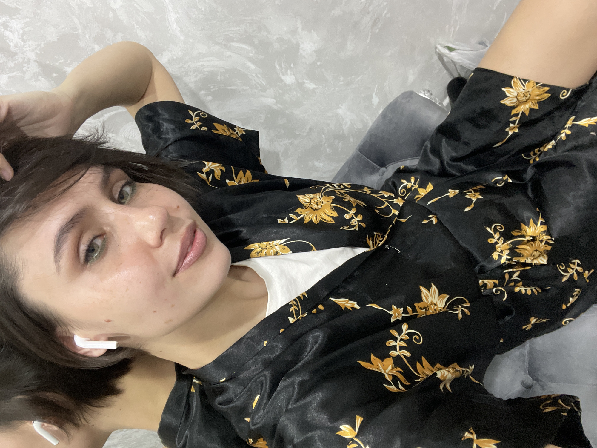 A female subject is depicted in an indoor setting, likely a private residence in Izhevsk, Russia. She has short, dark brown hair, green eyes, and fair skin with visible freckles. She is wearing a black satin robe adorned with yellow floral patterns over a white t-shirt. White wireless earbuds are present in both ears. Her right arm is raised, with the hand positioned near the back of her head. She is seated on a grey upholstered chair or sofa, partially visible in the lower frame. The background consists of a light grey wall with a textured, possibly stucco finish. A mirror on the left side reflects the back of the subject's head and neck. A white plastic bag is partially visible on the lower right of the frame. The subject is looking directly at the camera with a neutral expression. The precise location within Izhevsk, Russia, cannot be determined from the visual information provided.