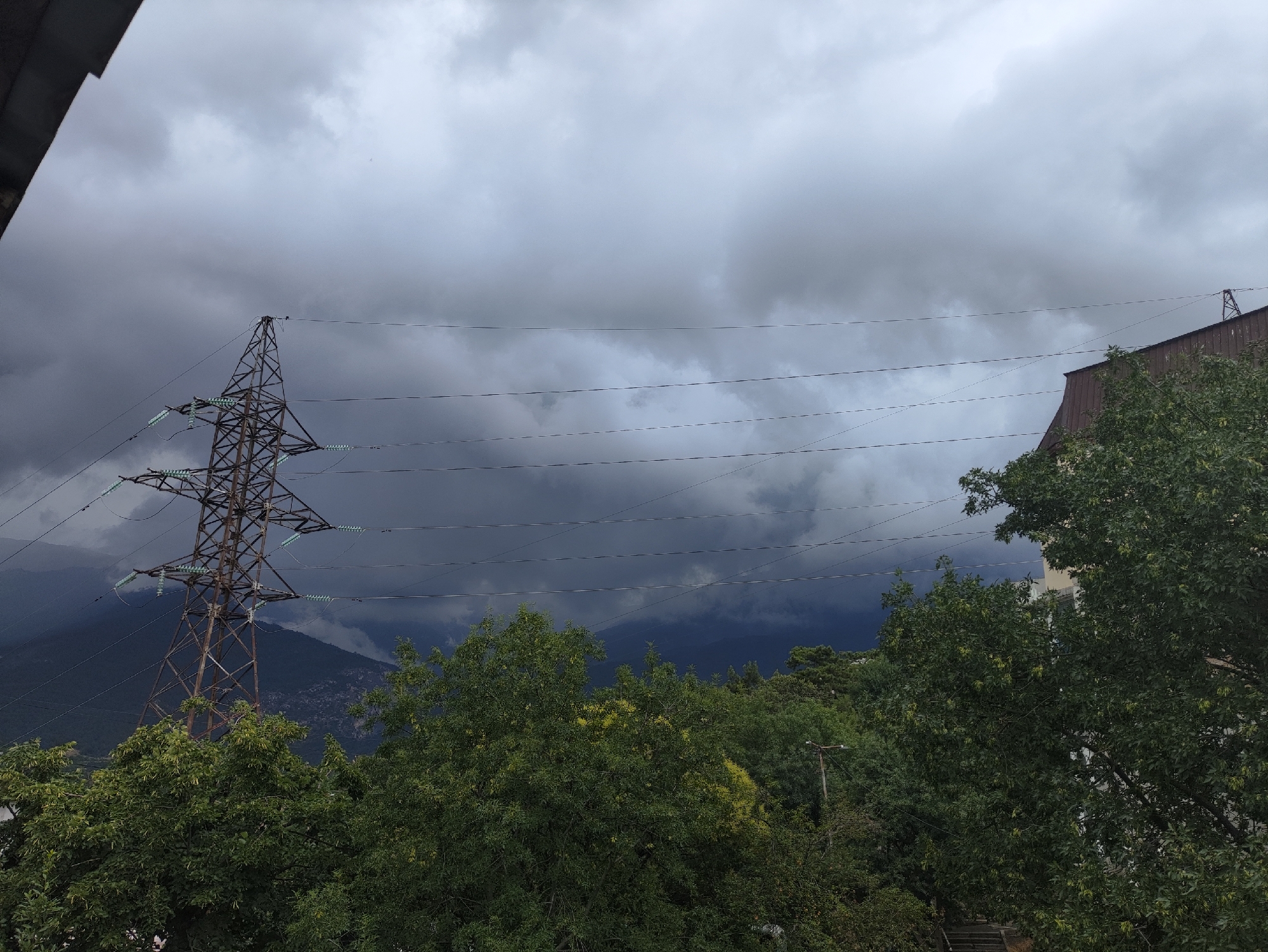 The image captures a dramatic sky over a mountainous landscape in Массандровский поселковый совет, Ukraine. Dark, heavy clouds dominate the upper portion of the frame, hinting at an impending storm. In the foreground, a tall, lattice metal electricity pylon stands prominently, supporting multiple power lines that stretch across the scene. To the right, a section of a building with a brown, sloped roof is visible, partially obscured by lush green trees. The lower part of the image is filled with dense foliage of various trees, some with vibrant green leaves, others appearing more yellow-green. The background features dark, hazy mountains, adding depth and a sense of scale to the environment. The overall atmosphere is somber and slightly foreboding due to the heavy cloud cover.