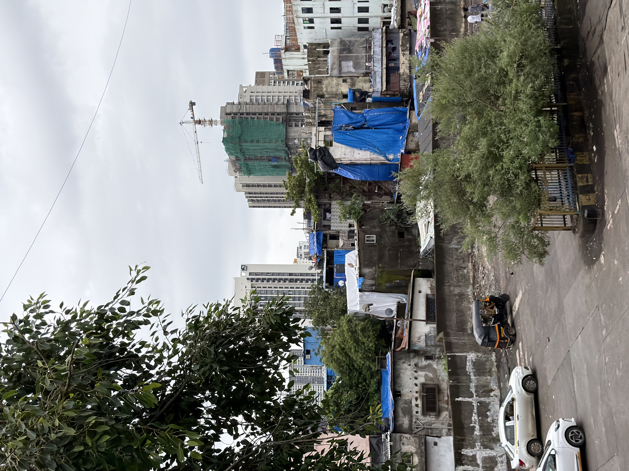 The image depicts a scene in the city of Mumbai, India. In the foreground, a paved road is visible with two white cars and a three-wheeled auto-rickshaw. A dense green tree with thin branches is situated on the right side of the frame, behind a metal fence. In the midground, there is a wall made of concrete and what appears to be a dense area of low-rise buildings with corrugated metal roofs and some covered with blue tarpaulins. Some of these structures show signs of wear and tear. Further back, several taller buildings, including modern apartment complexes, are visible. One of these taller buildings is under construction, wrapped in green netting, with a construction crane towering above it. The sky is overcast with clouds. The scene suggests a juxtaposition of urban development and older, more densely populated settlements within Mumbai.