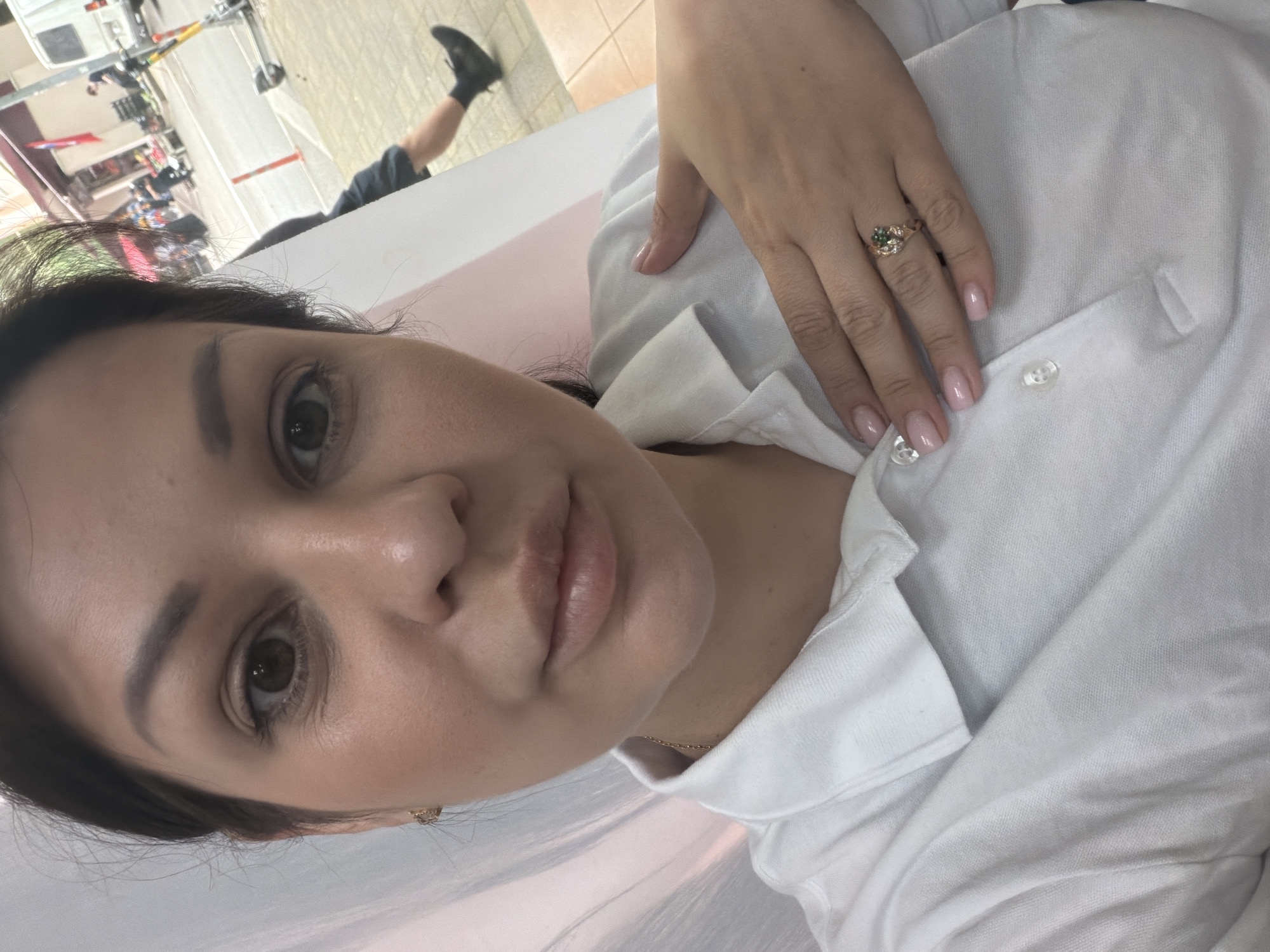 A woman, facing the camera, is taking a selfie in what appears to be a public area. She is wearing a white collared shirt and has her left hand placed on her chest, displaying a ring on her finger. In the background, a street scene is visible with a white van, a scooter, and several people. The presence of a Turkish flag in the background suggests the location might be near a market or a business that has ties to Turkey. Given the style of the architecture and the general ambiance, it is possible this scene is located in Sochi, Russia, specifically in an area with a mix of commercial activity and pedestrian traffic. The lighting is bright, indicating it is daytime.