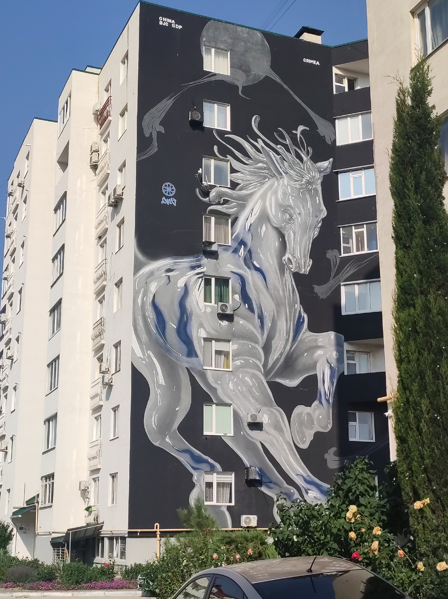This media file captures a mural on the side of a multi-story building. The mural features a large, stylized white horse with flowing mane and tail, depicted in motion against a black background. The horse appears translucent, with blue and grey shading giving it a dynamic, almost ethereal quality. The mural covers a significant portion of the building's facade, with windows of the building integrated into the artwork.
The building itself is a light-colored, multi-story apartment block with numerous windows and balconies. To the right of the mural, a tall, dark green evergreen tree stands, partially obscuring the edge of the building. Below the mural, at street level, there is greenery including flowering bushes with yellow and pink blooms, and a paved area. A car is partially visible in the foreground, suggesting this is a street-level view.
Visible text in the image includes "CHINA BJC GDP" and "CRIMEA" written in white at the top of the mural. There is also a circular emblem with intricate lines and what appears to be the letters "AWG" beneath it.
The scene is illuminated by bright sunlight, indicating a clear day. The shadows and the intensity of the light suggest it is daytime, likely late morning or afternoon. The overall mood conveyed by the mural is one of power and movement, contrasting with the stillness of the urban setting.
