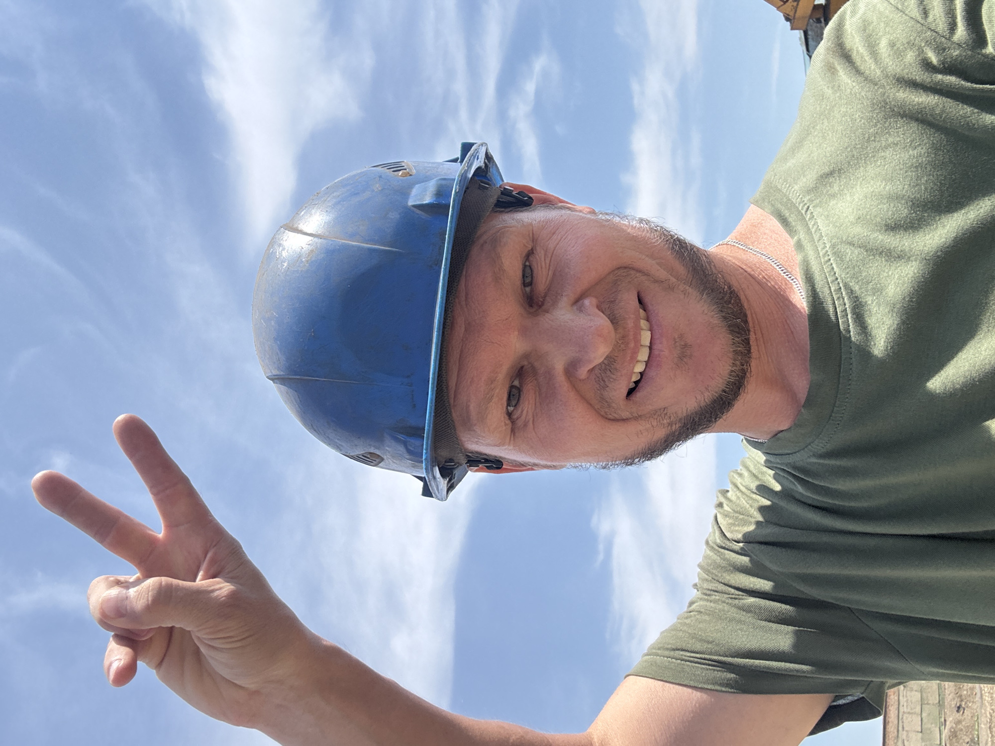 A man wearing a blue hard hat and a green t-shirt smiles at the camera while holding up two fingers in a peace sign. He has a beard and is outdoors with a blue sky and clouds in the background. The image was taken in Oskemen, Kazakhstan.