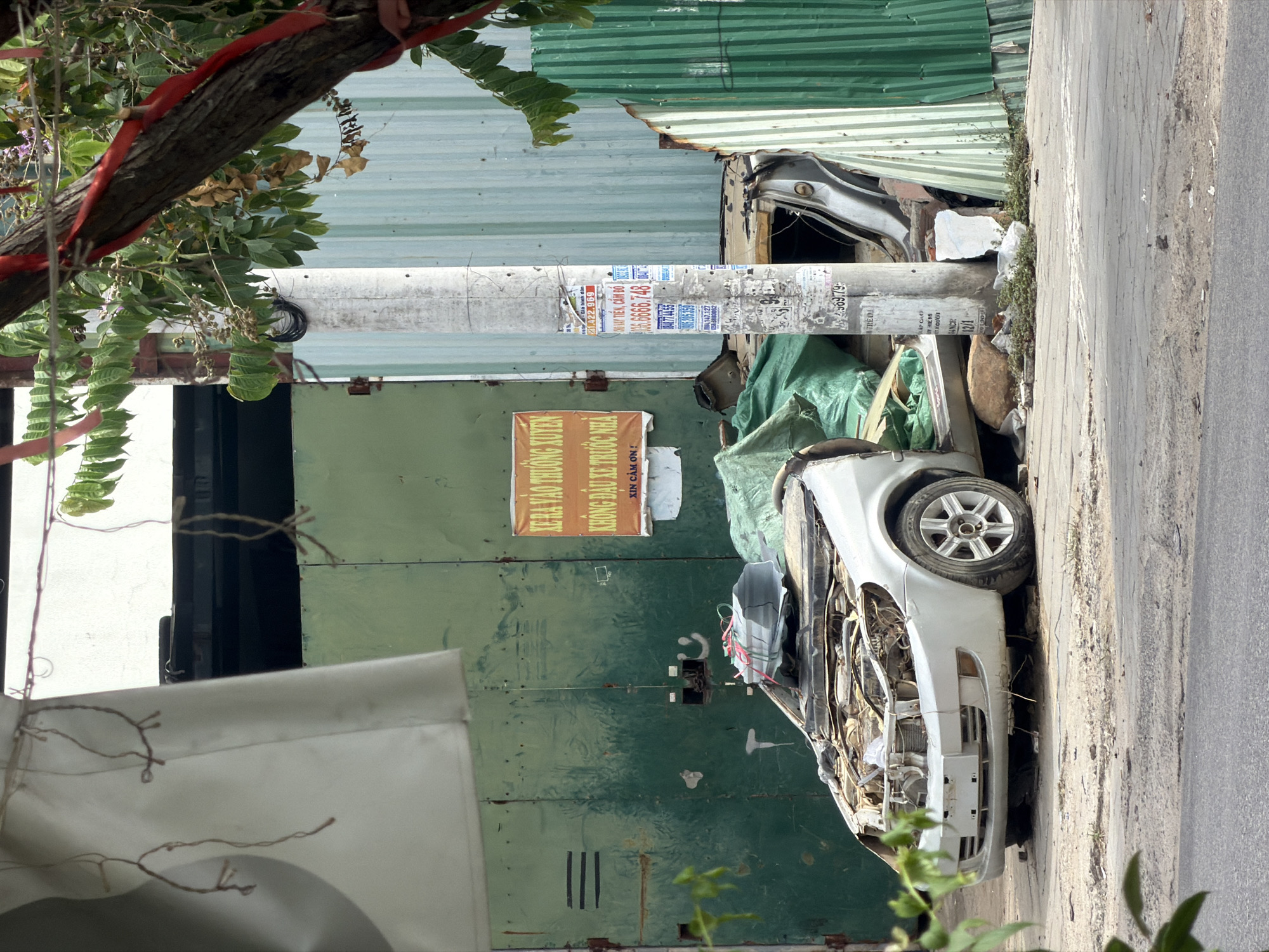 A partially dismantled, white sedan sits on a dirt and asphalt surface in front of a green corrugated metal fence. The car's hood is open, revealing exposed engine components, and its front bumper and grille are intact. A green tarp is draped over the engine area, and other debris, including rocks and greenery, are scattered around the vehicle. A white utility pole stands to the right of the car, adorned with various posters and flyers, some of which are in Vietnamese. To the left of the car, a section of a green metal garage door is visible. The background consists of the corrugated metal fence and a portion of a building. Red ribbons are tied to a tree branch in the upper right of the frame, and a white tarp hangs on a wire in the upper left. The scene appears to be an outdoor area in a city, possibly a junkyard or an informal repair site. The presence of Vietnamese text on the signs and the general urban environment suggest the location is in Nha Trang, Vietnam.