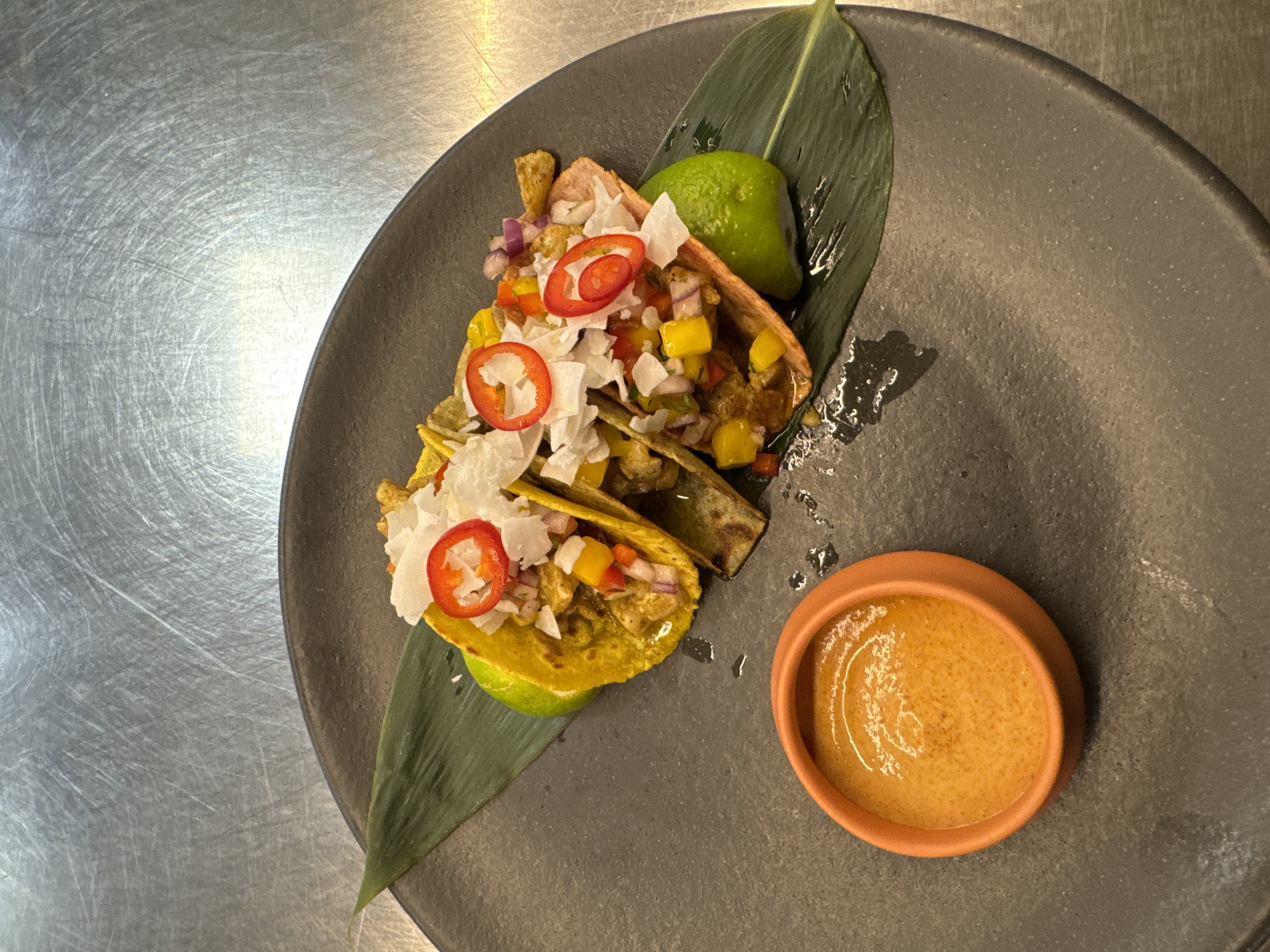 A close-up, overhead shot captures a plate of three tacos resting on a large green leaf. The tacos are small and appear to be filled with a savory mixture topped with shredded white cheese, diced red peppers, and chili slices. A small, round, orange dish containing a creamy, orange sauce sits to the left of the tacos. The plate is dark and textured, contrasting with the vibrant colors of the food. The background is a metallic, scratched surface, likely a countertop. This image was taken in Sofia, Bulgaria.