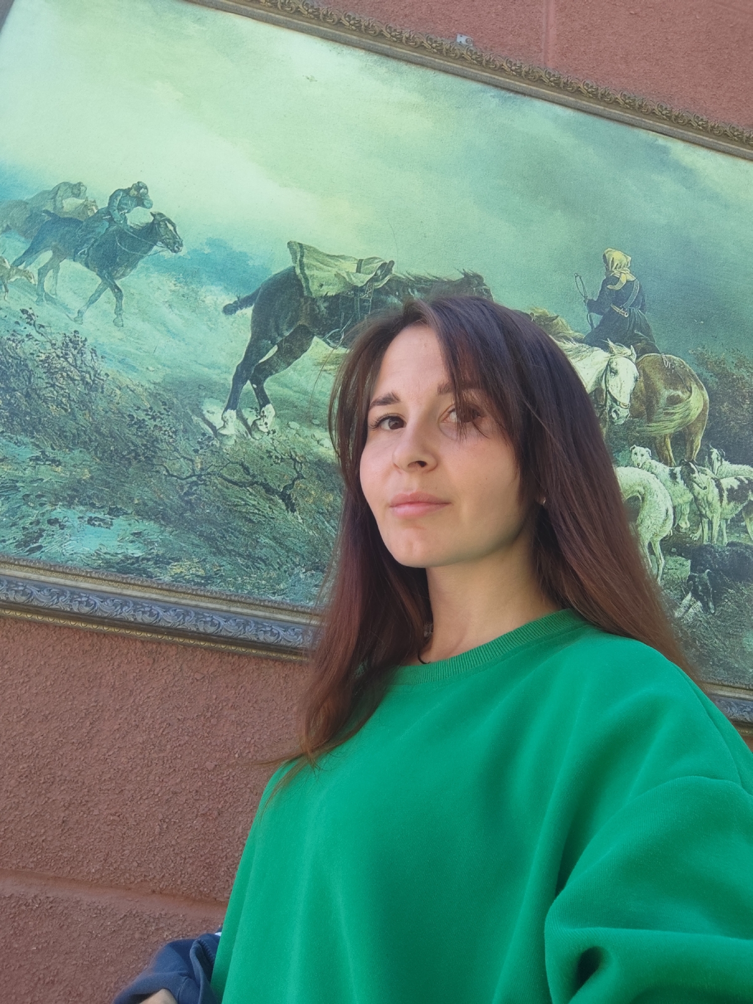 A young adult female stands in the foreground, positioned slightly to the left of the frame's center. She has long, dark brown hair, parted slightly off-center, falling over her shoulders. Her gaze is directed towards the camera with a neutral expression and a slight head tilt. She wears an emerald green crew-neck sweatshirt. A dark blue garment is partially visible, held in her left hand, which is mostly cropped from view.
Behind her is a large, rectangular, framed artwork affixed to an exterior wall in Chaikovsky, Russia. The artwork is framed by an ornate, dark gold-colored border with intricate scrollwork. The painting depicts a dynamic outdoor scene with a greenish-blue dominant hue. On the left side, multiple horses and riders are shown in motion, one dark horse prominently featured with a jockey in a cap and dark attire. The ground beneath them is a dark, overgrown landscape. On the right side, a figure cloaked in a hooded garment, possibly a monk or shepherd, is seated on a light-colored horse, viewed from behind. Below this horse and rider is a gathering of several sheep and possibly goats, predominantly white with some darker markings. The sky in the upper portion of the painting is a hazy, pale blue-green. The painting appears somewhat aged or weathered.
The background wall, visible above and below the painting, has a coarse, textured, reddish-brown or terracotta surface, suggesting stucco or aggregate. The scene is illuminated by natural light, indicating daytime.