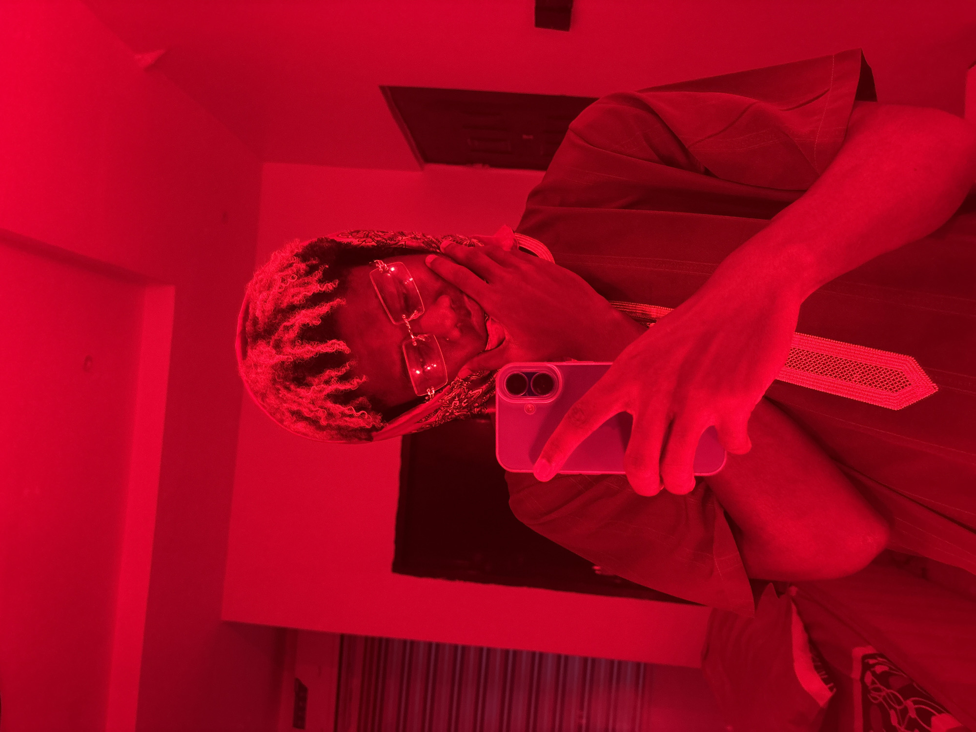 An individual is captured in a selfie taken indoors in Igboefon, Nigeria, under pervasive red lighting. The subject, positioned centrally, is observed from the chest up, with their head slightly tilted downwards. They possess lighter-toned dreadlocks or styled hair and wear clear, rectangular-framed glasses. A patterned fabric, possibly a bandana, is draped around their neck and partially over their head. The individual is wearing a dark-colored garment, potentially a tunic, featuring a vertical patterned strip down the front. Their right hand holds a light-colored smartphone vertically, with the dual camera lenses visible on the rear, indicating a mirror selfie or use of the front-facing camera. Their left hand rests gently against their chin or lower cheek. The background reveals a room with plain walls, an air conditioning unit on the left, a dark door on the right, and soft furnishings (pillows) partially visible in the lower-left corner. The entire scene is uniformly saturated in red light.